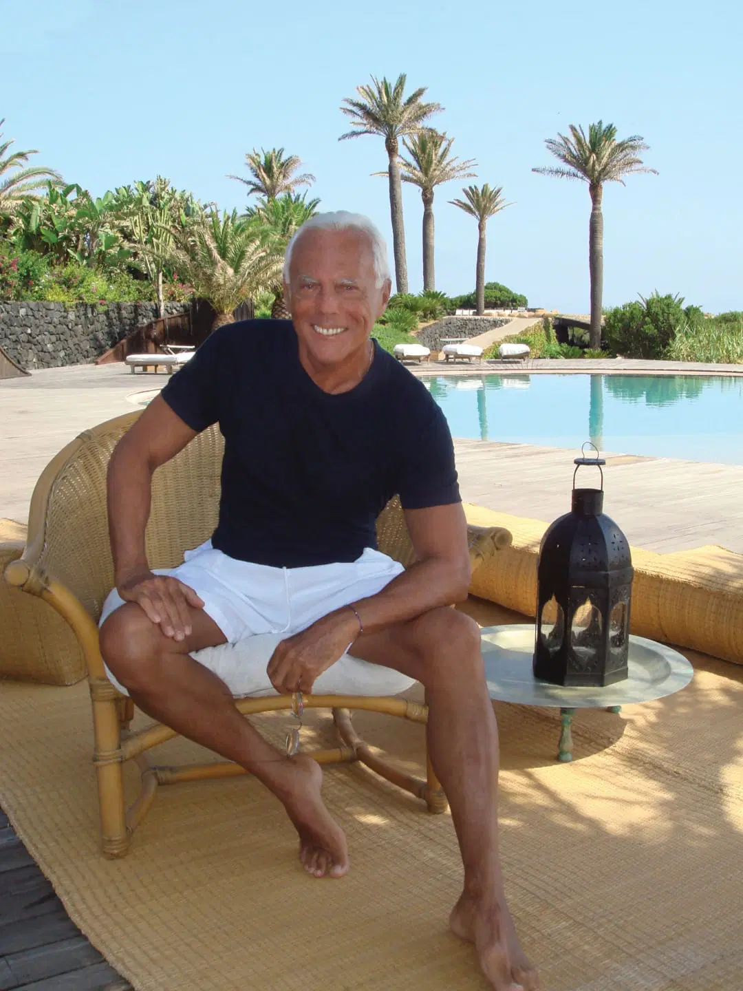 Giorgio Armani seduto su una poltroncina in vimini a bordo piscina, con una lanterna in primo piano