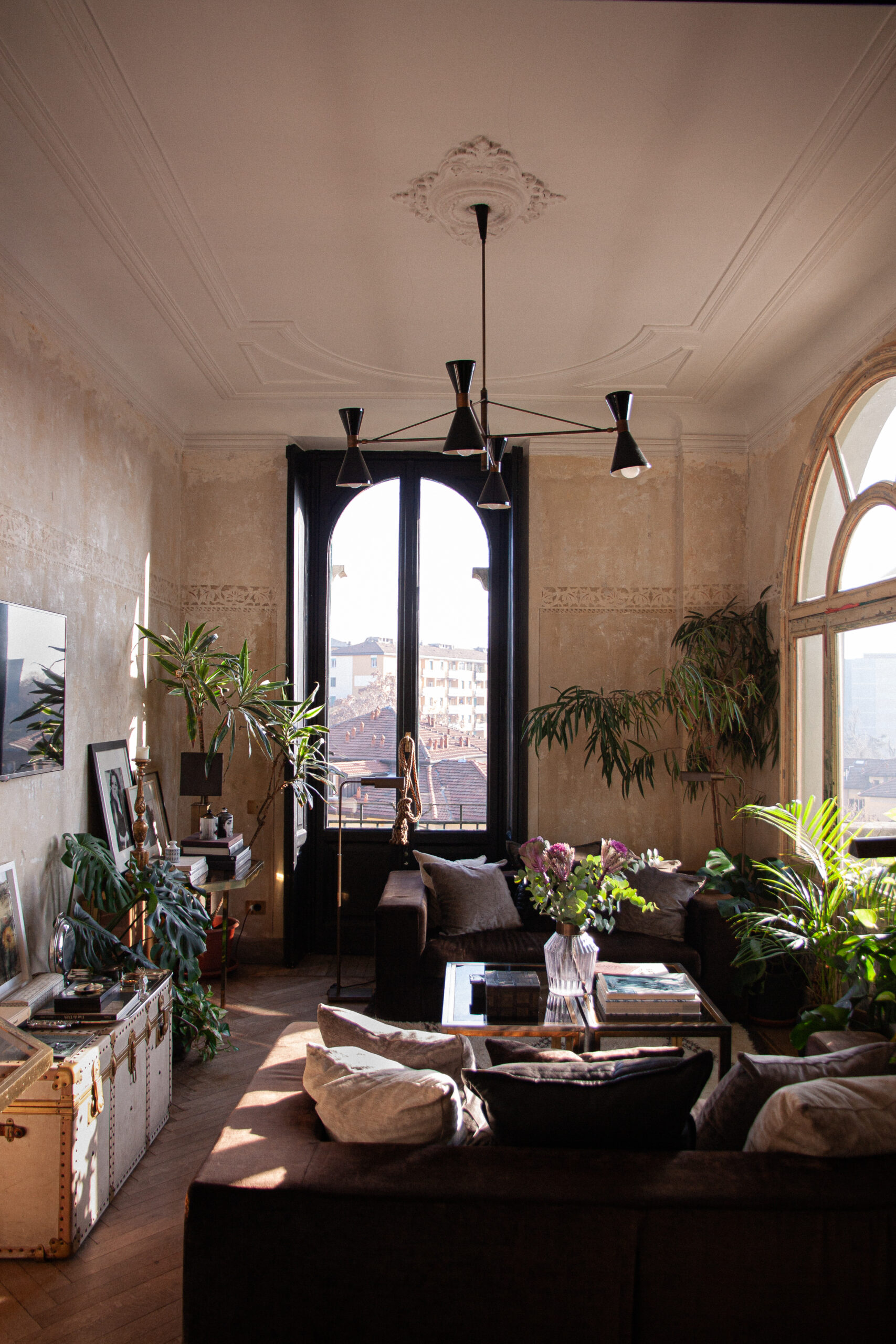 Soggiorno con soffitto alto e decorato, due finestre ad arco, divani bruni, tavolino in vetro e numerose piante.