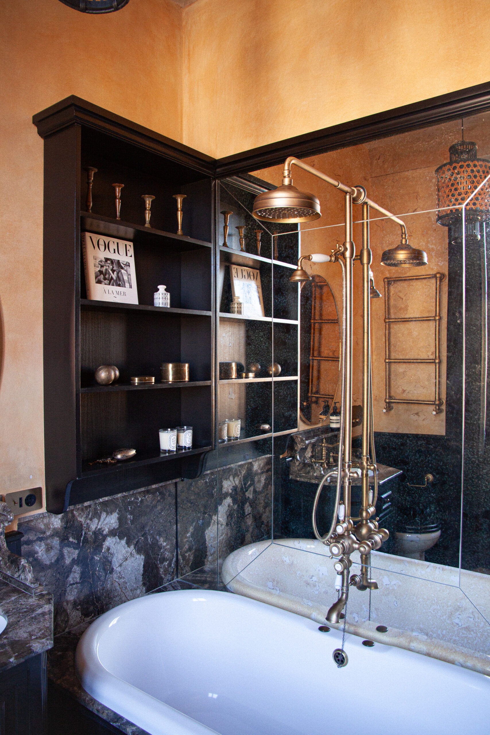Bagno con vasca bianca, doccia e rubinetteria color ottone, rivestimenti scuri venati, scaffali e pareti color ocra.