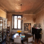 Sala da pranzo con tavolo rotondo scuro, sedie imbottite, vetrina, lampadario color ottone e pareti nei toni della terra.