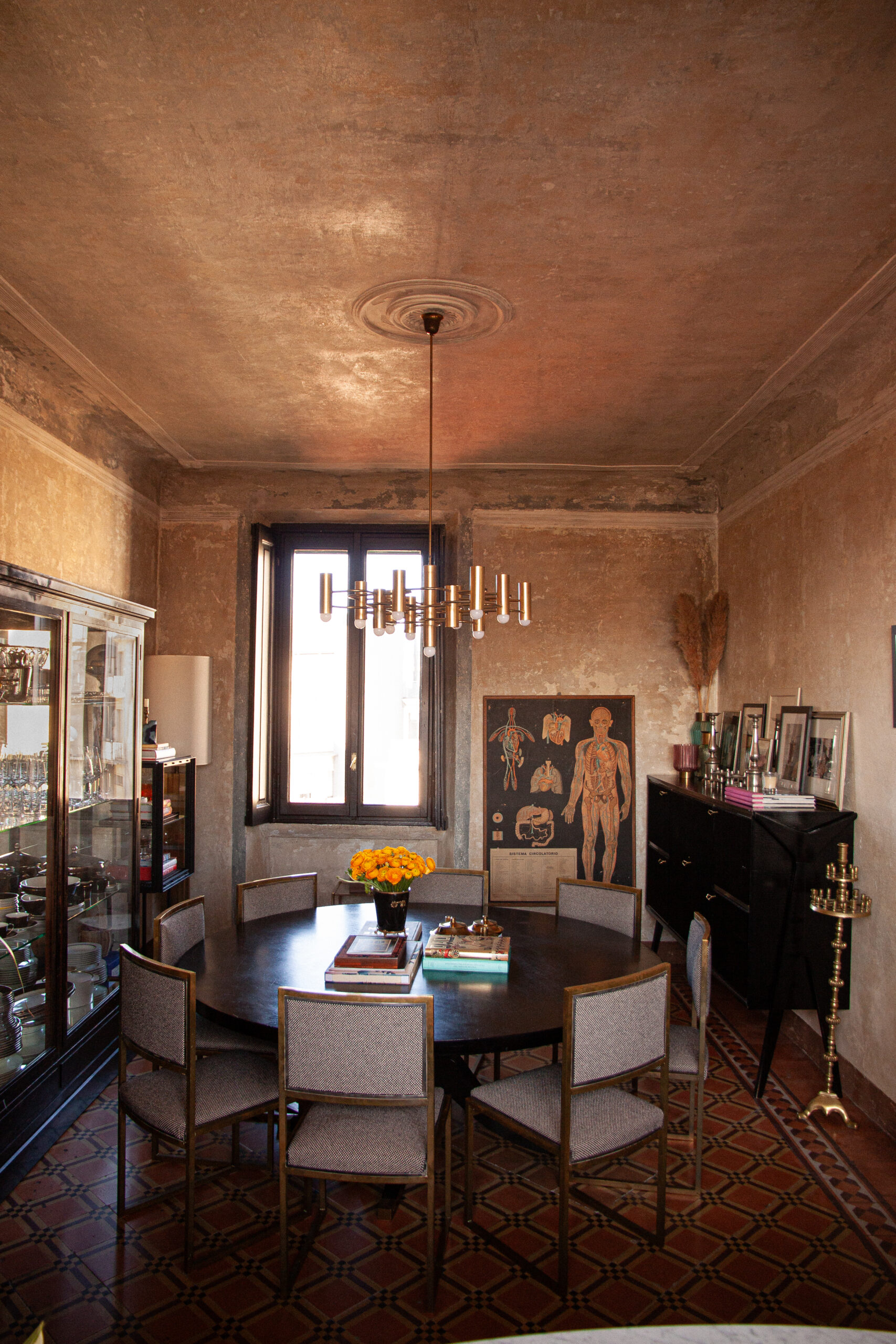 Sala da pranzo con tavolo rotondo scuro, sedie imbottite, vetrina, lampadario color ottone e pareti nei toni della terra.