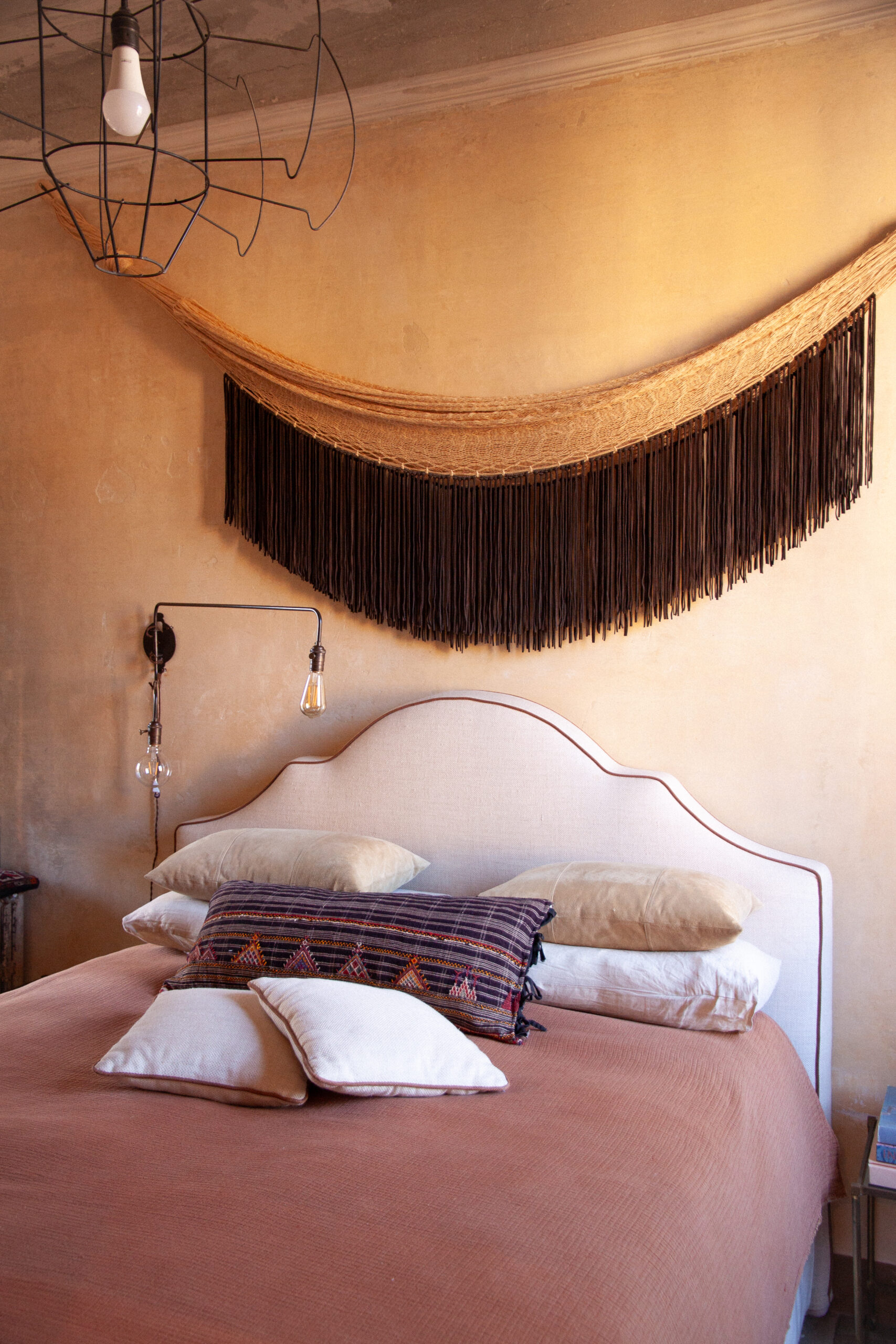 Letto con copriletto rosa, testiera chiara sagomata e cuscini, sotto una decorazione tessile a frange su parete color ocra.