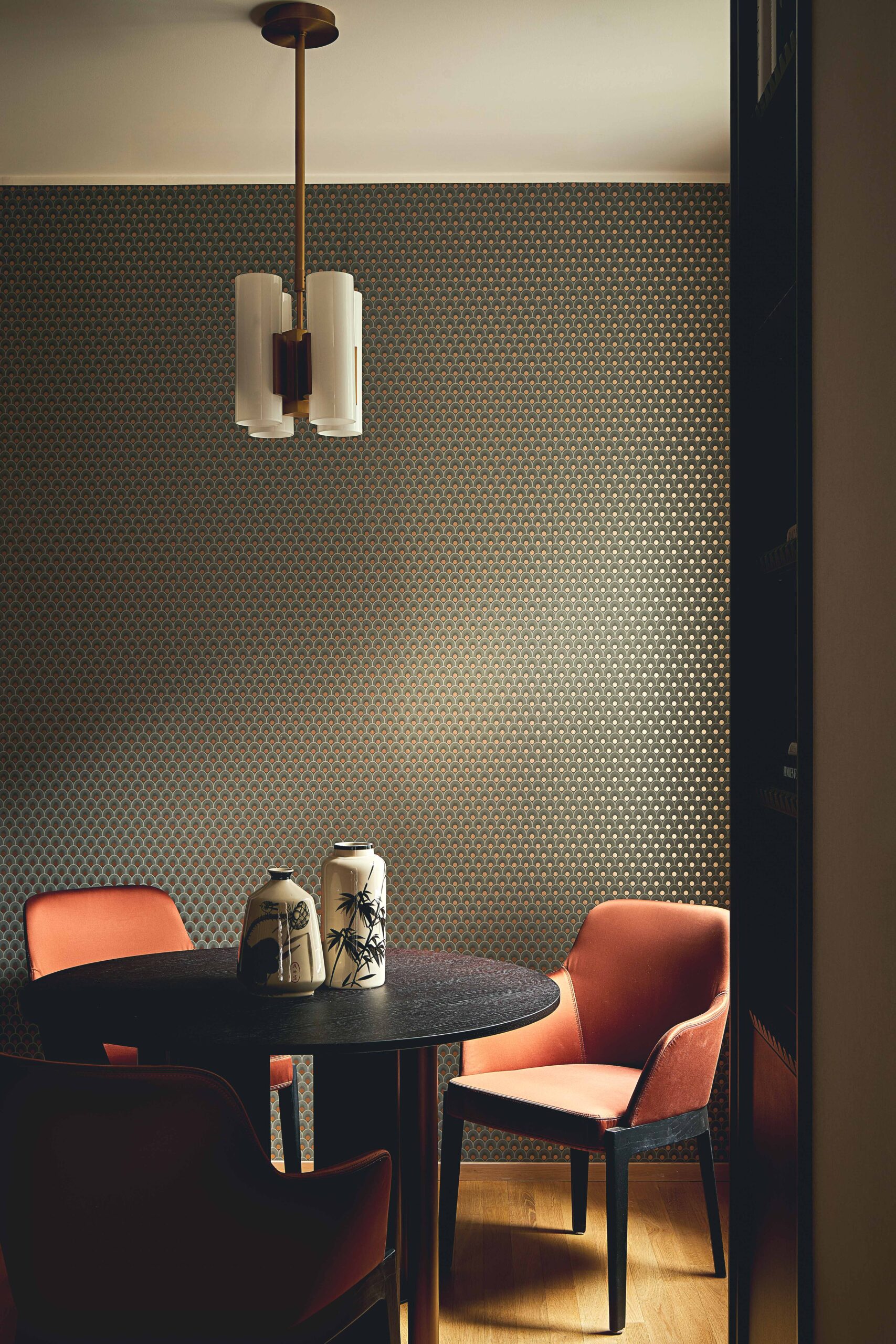Tavolo da pranzo scuro con sedie color cuoio, lampada a sospensione e carta da parati a piccoli motivi geometrici.