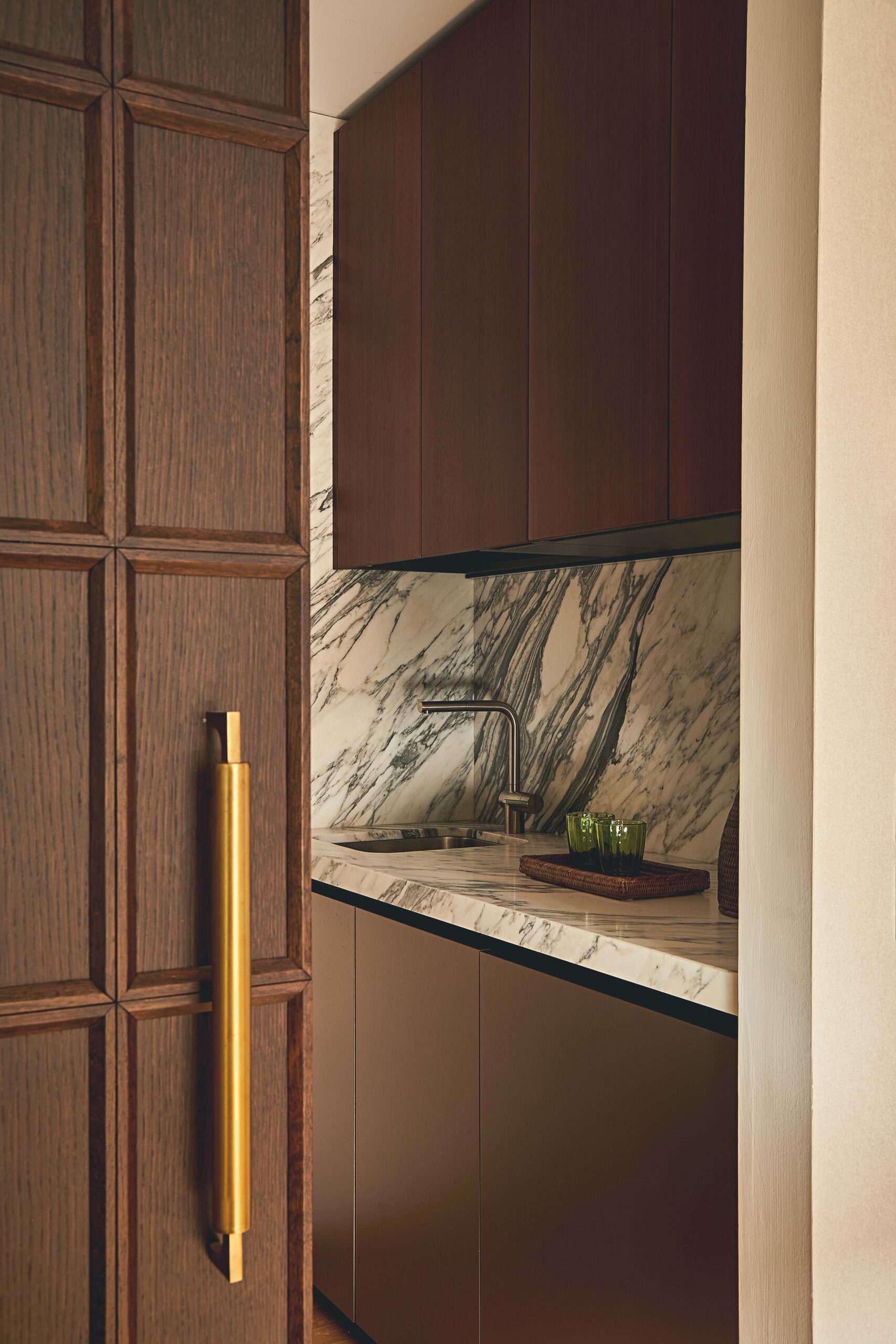 Porta scorrevole in legno con maniglione in ottone davanti a una cucina con pensili scuri e piano in marmo venato.