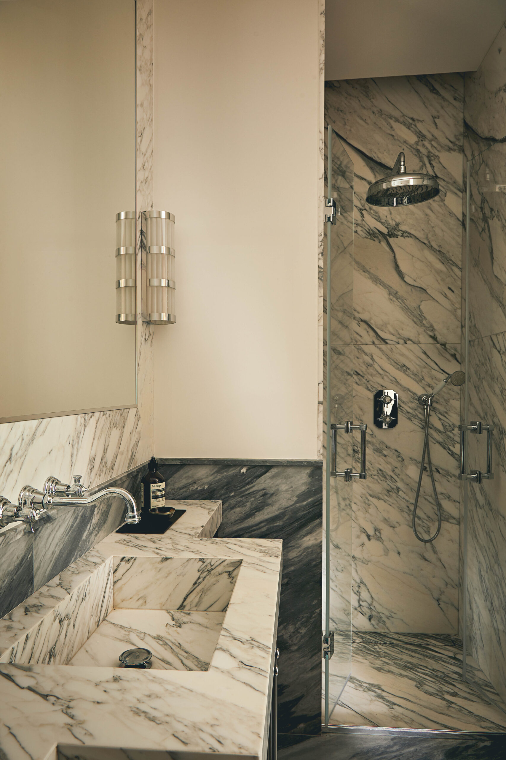 Bagno con lavabo integrato in marmo venato, specchio, applique cilindrica e doccia rivestita in marmo.