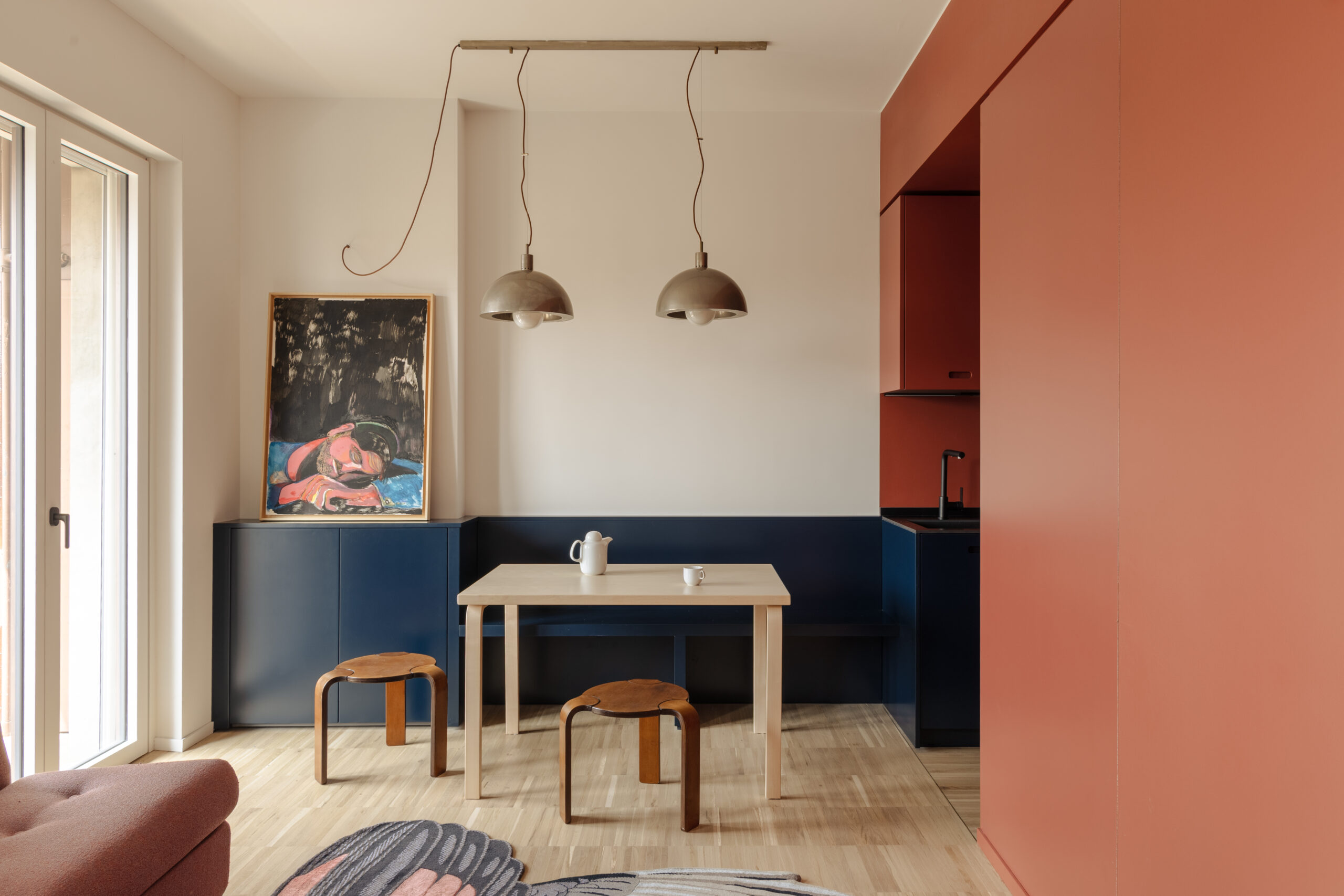 Monolocale di THAT Z SHOW con tavolo chiaro, sgabelli in legno, panca blu e volume cucina rosso terracotta.