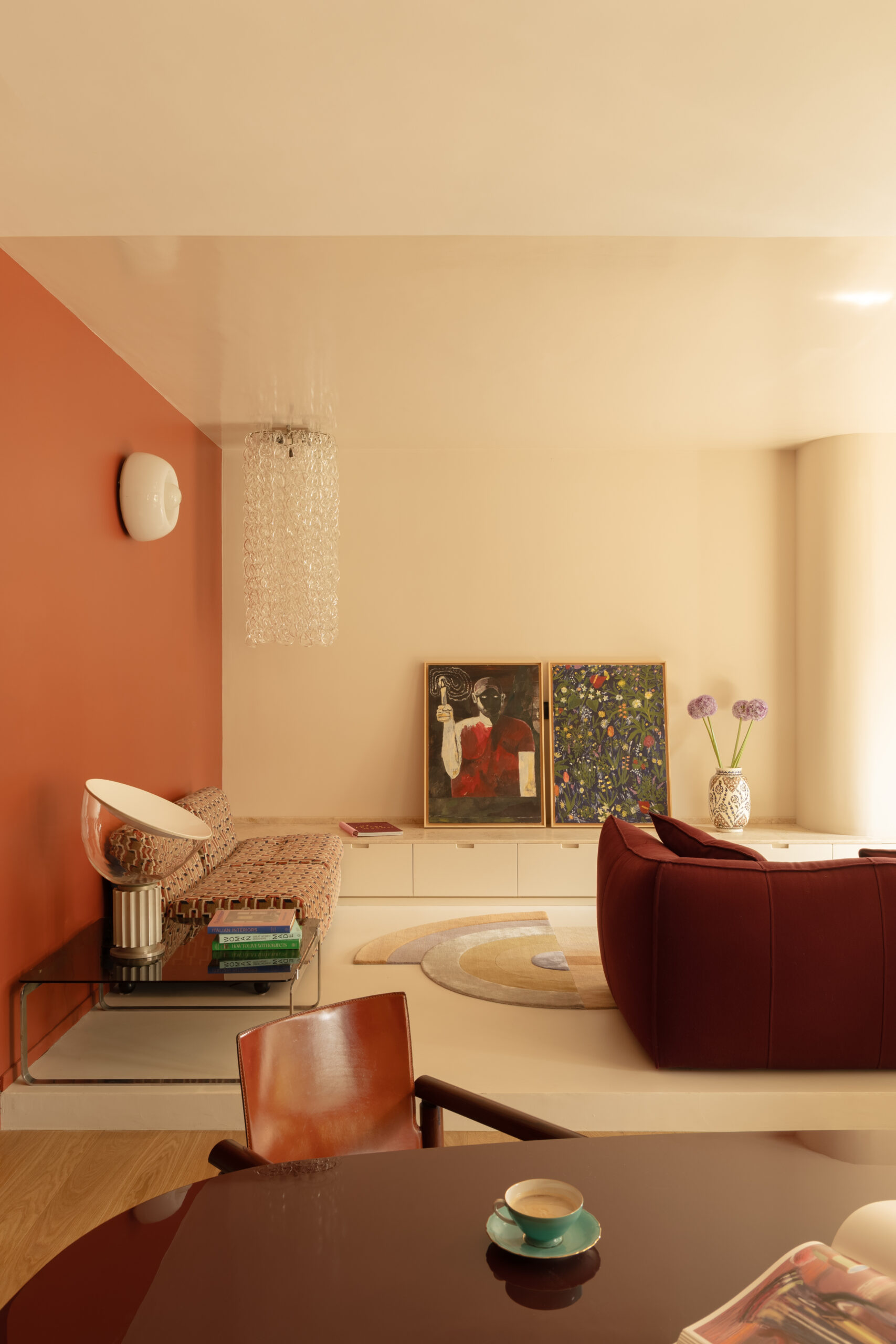 Soggiorno rialzato con divano bordeaux, seduta a motivi geometrici, opere appoggiate alla parete e lampada in vetro.