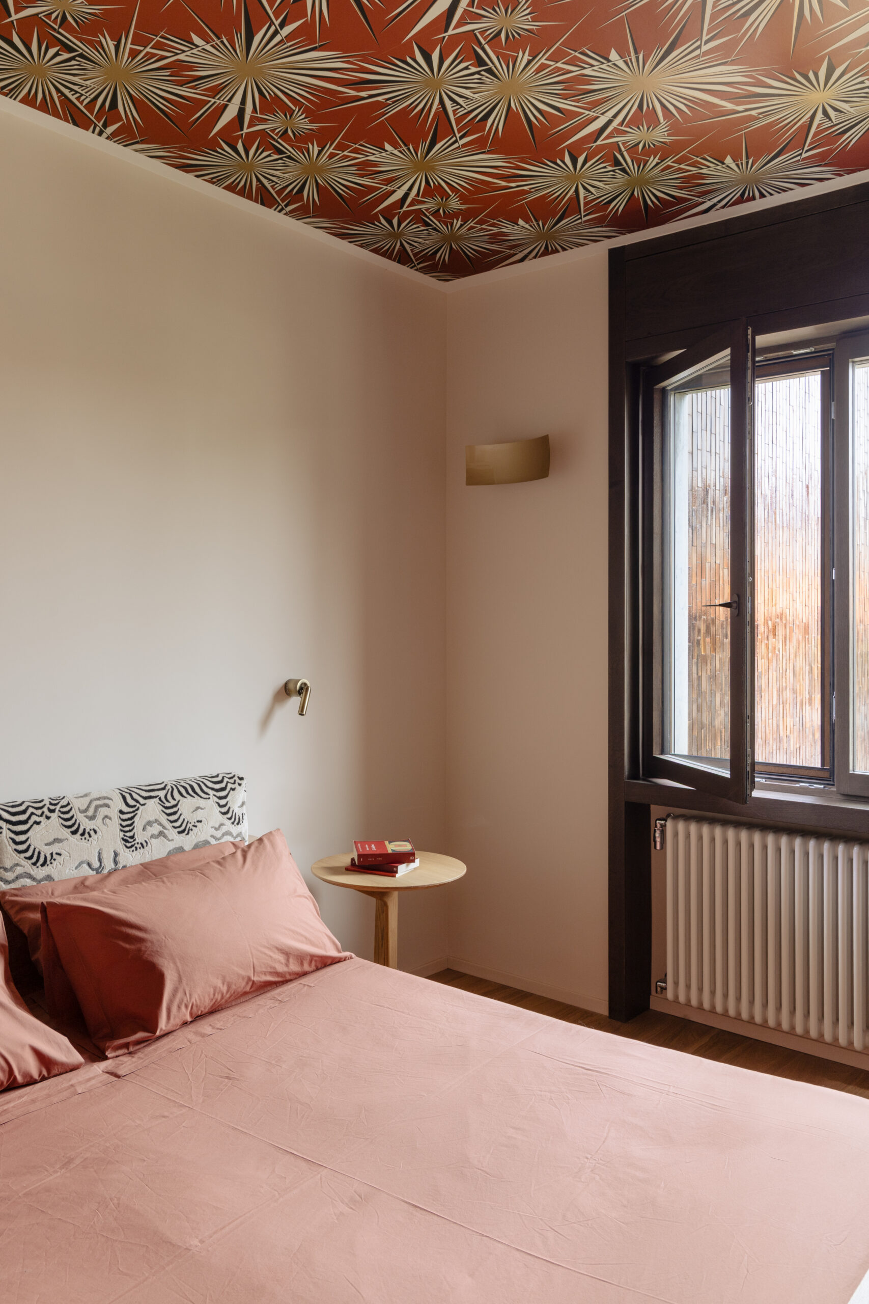 Camera con biancheria rosa, testata a motivi neri e bianchi, soffitto decorato e finestra con cornice scura.