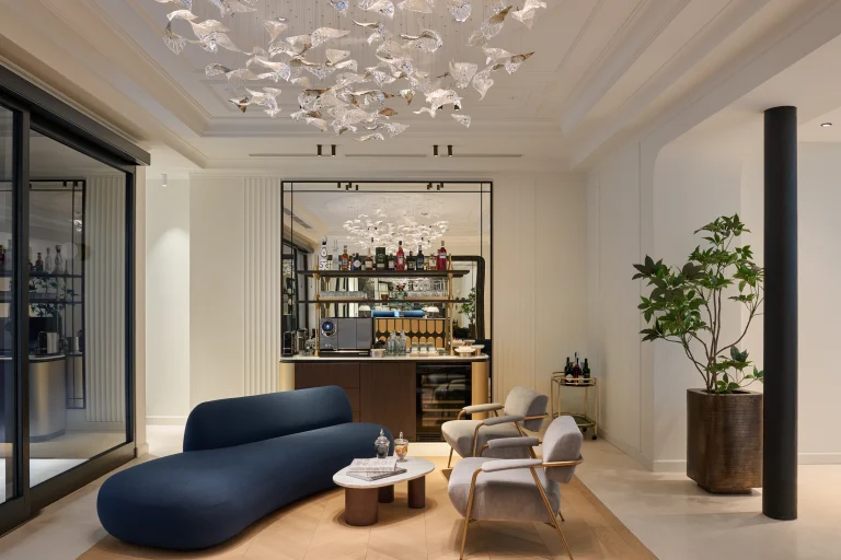 Lobby con divano blu curvo, poltrone chiare, tavolini e lampadario bianco, aperta verso il bar.