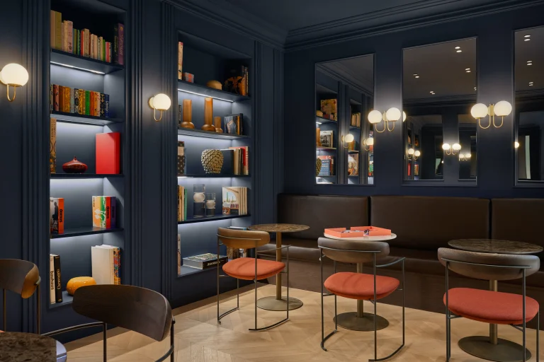 Sala con librerie blu, panche scure, tavoli rotondi, sedie rossastre e piccole lampade.