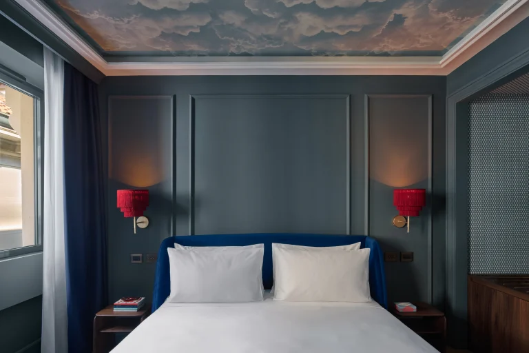 Letto con biancheria bianca tra due lampade rosse, pareti blu a riquadri e soffitto dipinto a nuvole.