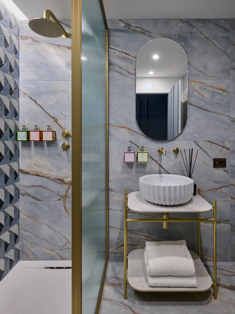 Bagno con lavabo bianco scanalato, specchio ovale, pareti venate, doccia e struttura metallica color ottone.