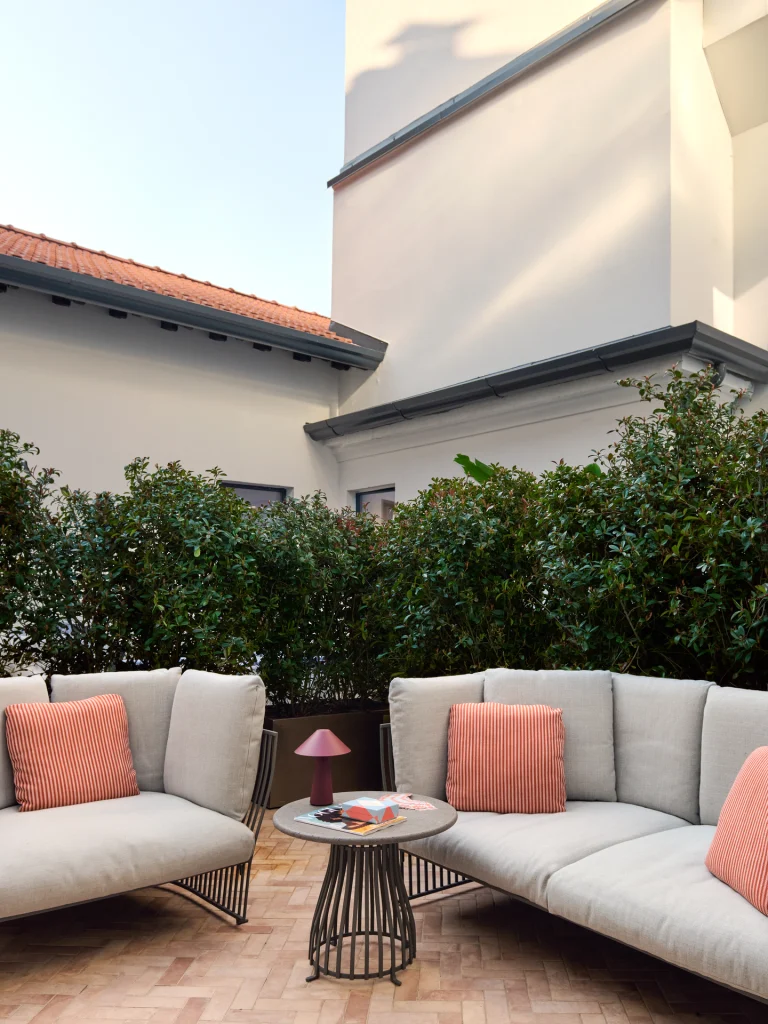 Terrazza con divano chiaro, cuscini rosa, tavolino rotondo e piante davanti a pareti chiare.