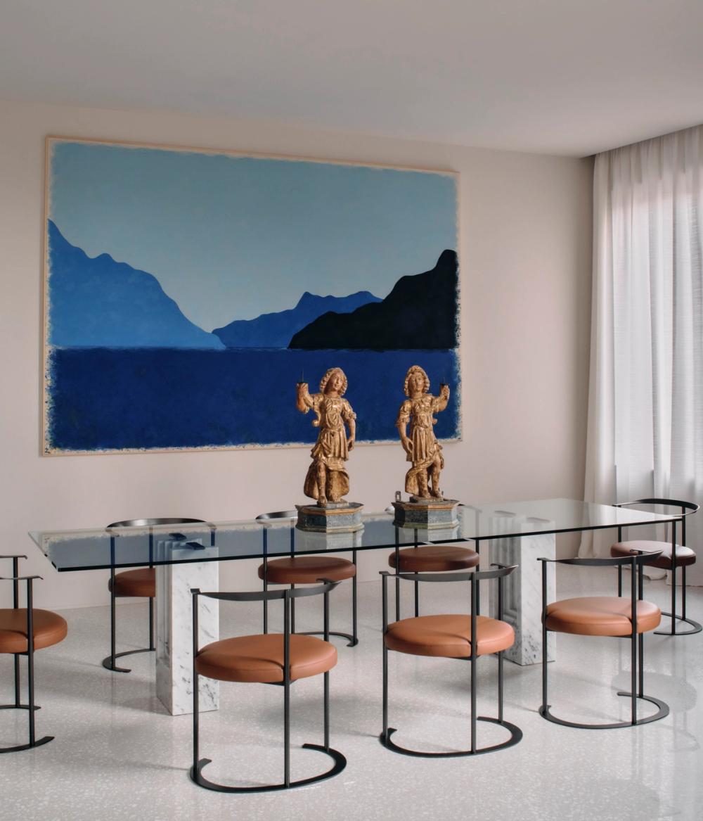 Sala da pranzo con tavolo in vetro, sedie metalliche con sedute color cuoio, due figure decorative e grande quadro azzurro.