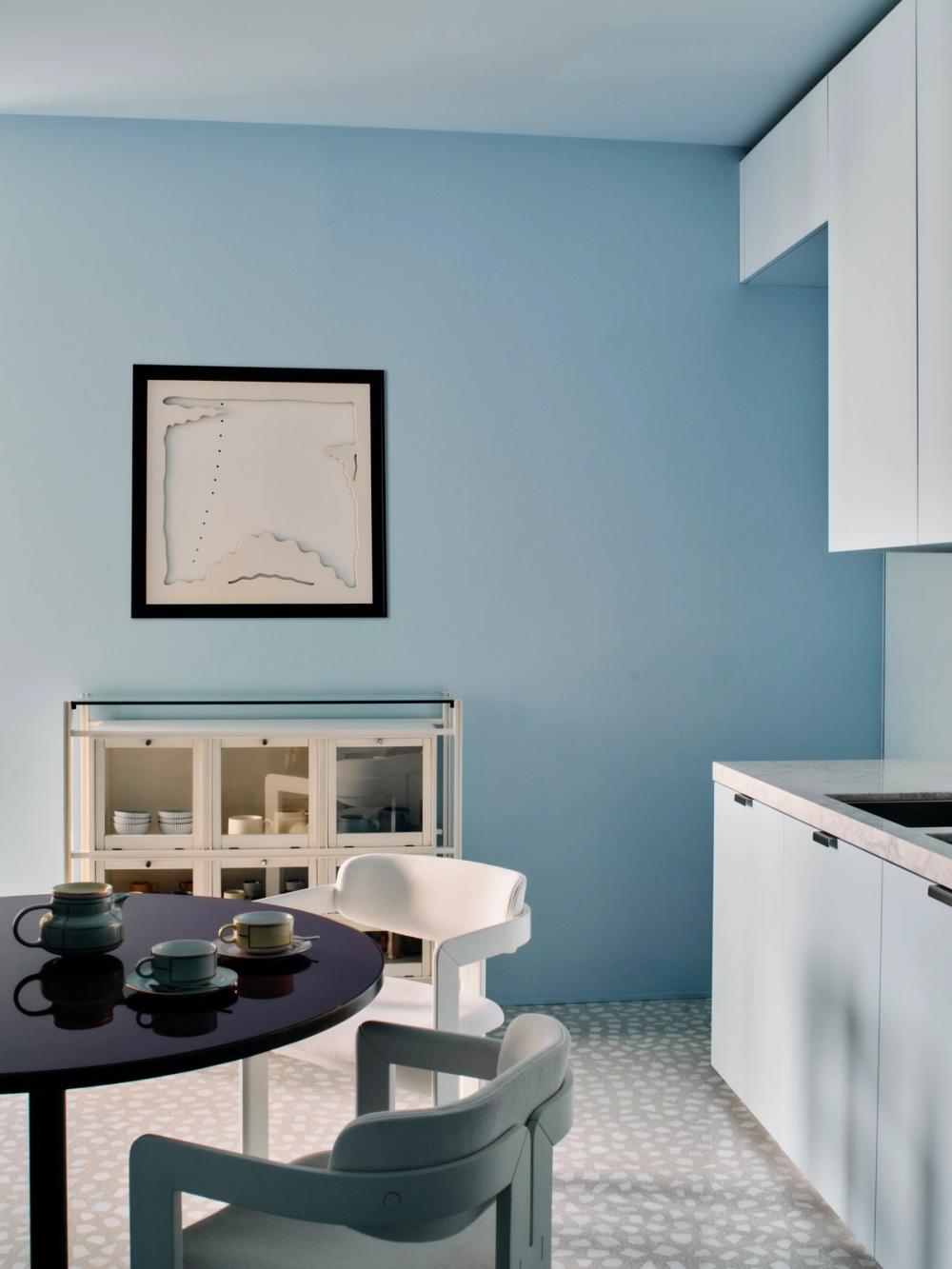 Cucina azzurra con mobili chiari, vetrina, tavolo scuro, sedie azzurre e pavimento a piccoli motivi.