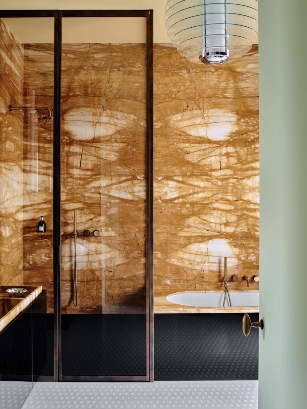 Bagno con rivestimento ambrato venato, doccia dai profili scuri, vasca bianca e pavimento a piccoli motivi geometrici.