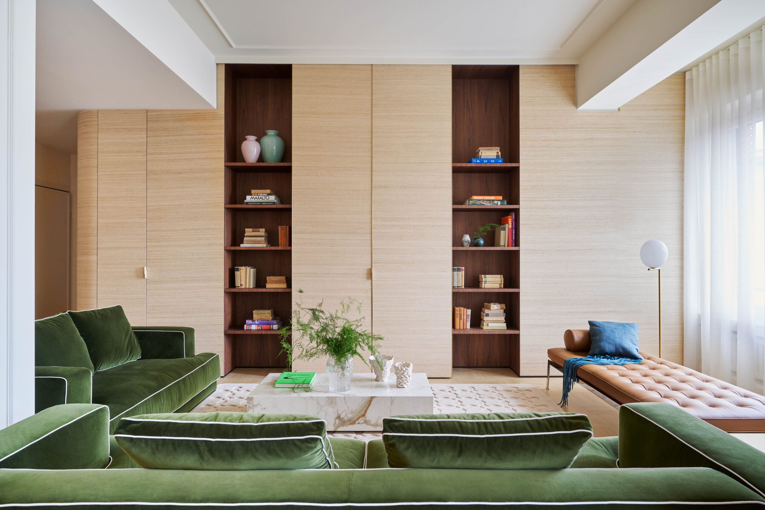 Soggiorno con divani verdi, tavolino in marmo, chaise longue in cuoio e due librerie verticali incassate nella parete chiara.