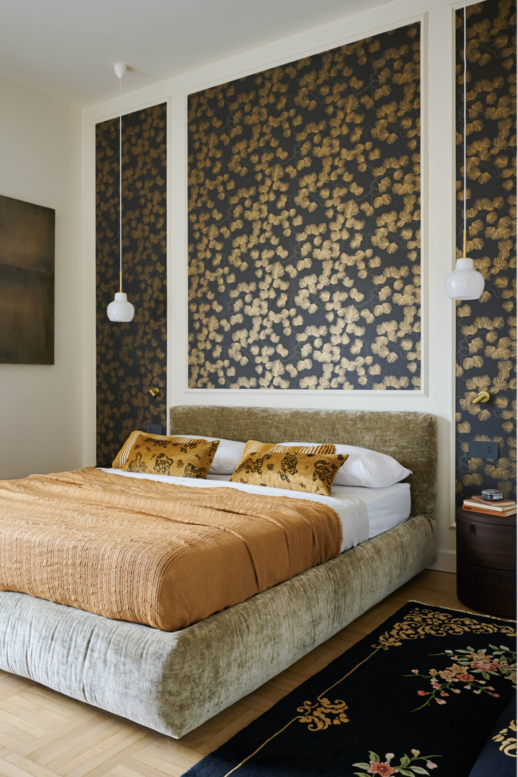 Letto imbottito chiaro con coperta ocra, parete scura a foglie dorate fra cornici bianche, due lampade sospese e tappeto floreale.