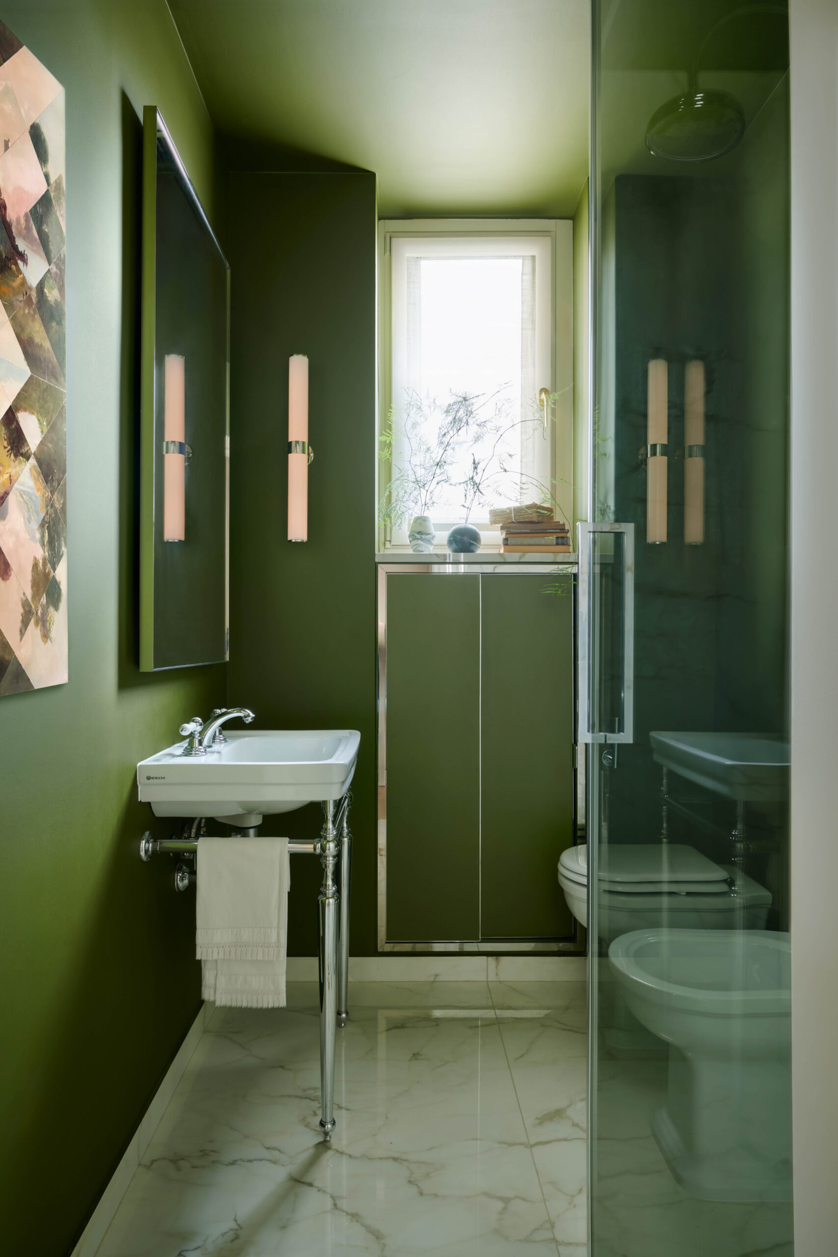 Bagno stretto con pareti e soffitto verdi, lavabo bianco su gambe cromate, specchio alto, doccia vetrata a destra e finestra sul fondo.