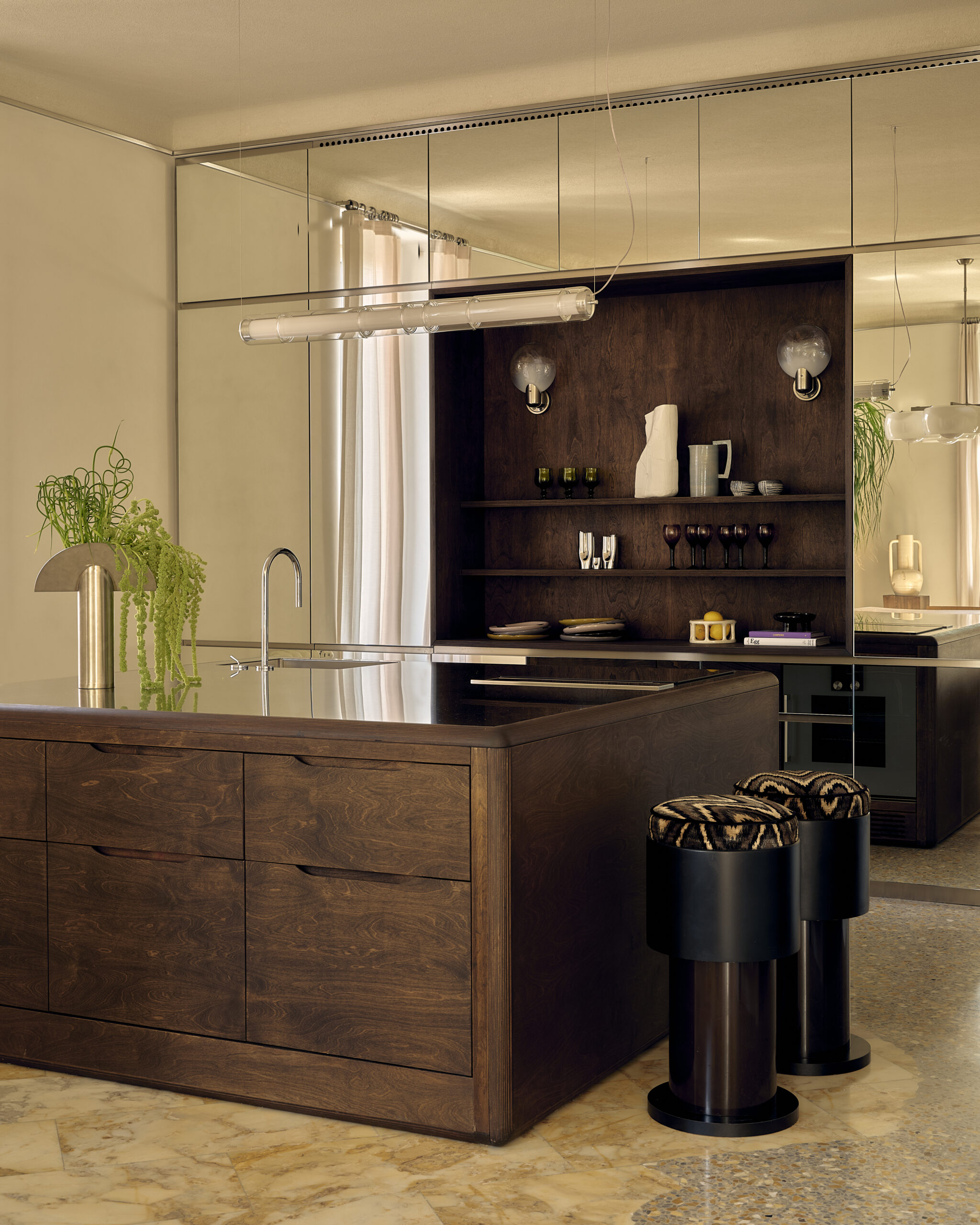 Cucina con isola in legno scuro, piano metallico, due sgabelli neri, nicchia con mensole e parete a specchio; marmo e seminato a pavimento.