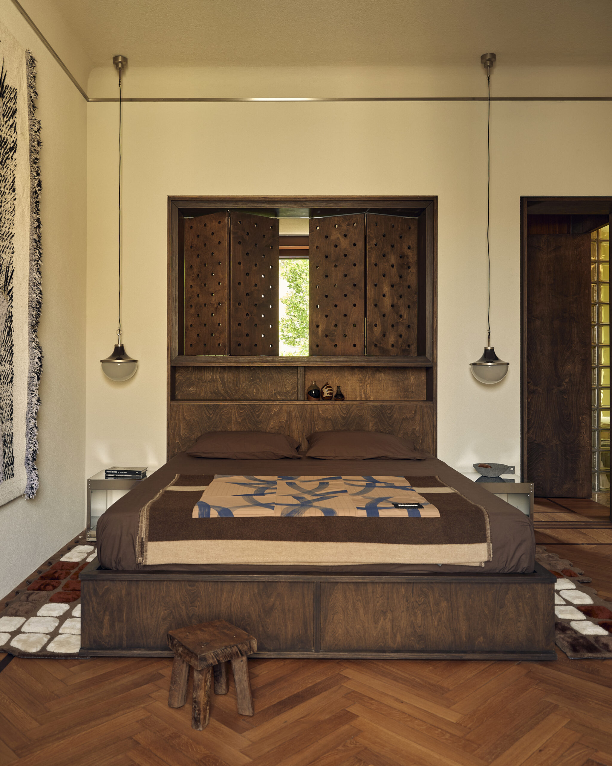 Camera con letto in legno scuro, tessili marroni, imposte traforate dietro la testata, due lampade sospese e parquet a spina di pesce.