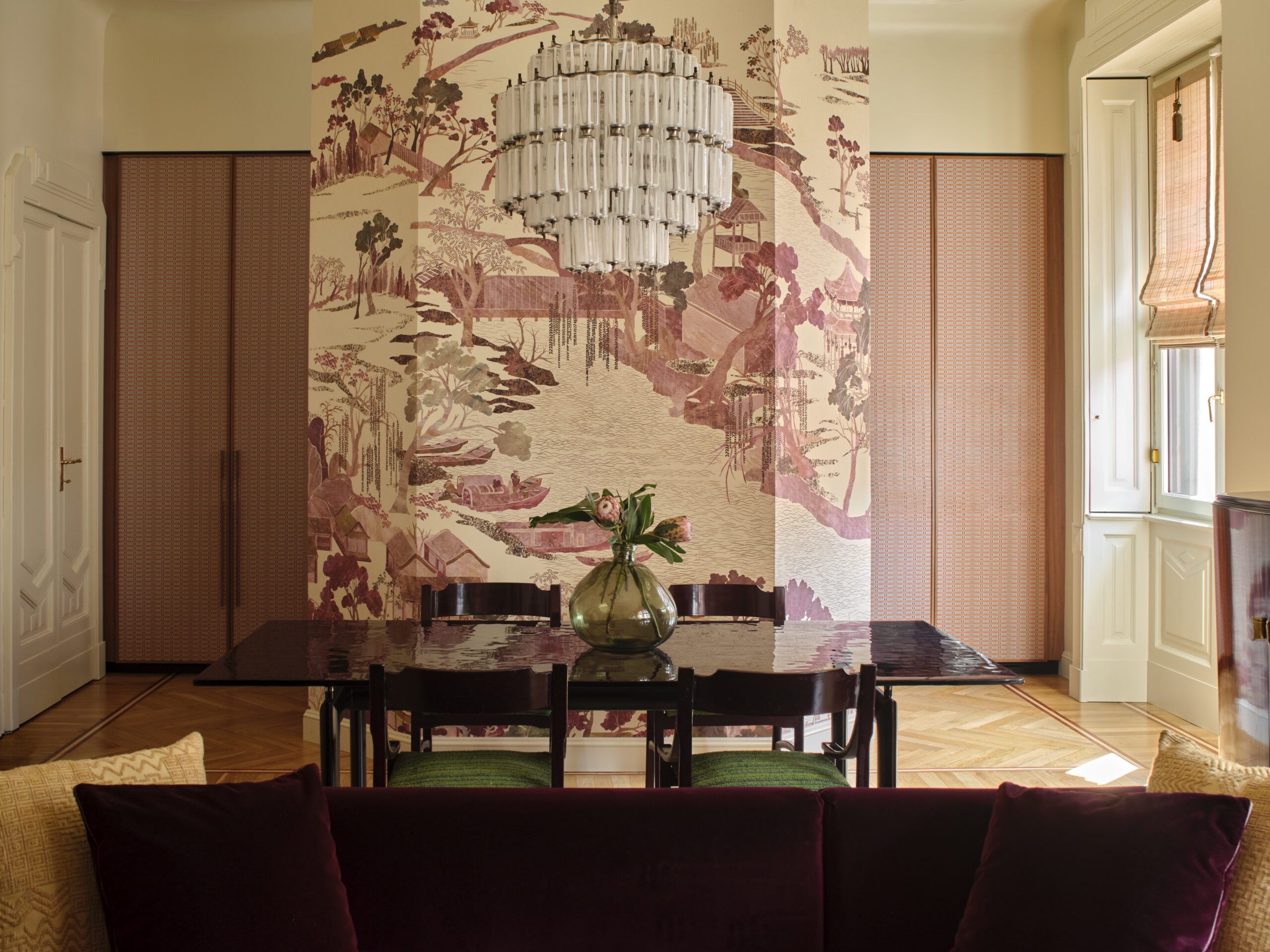 Zona pranzo con tavolo scuro, sedie verdi e lampadario in vetro davanti a una parete decorata con un paesaggio; divano color prugna in primo piano.