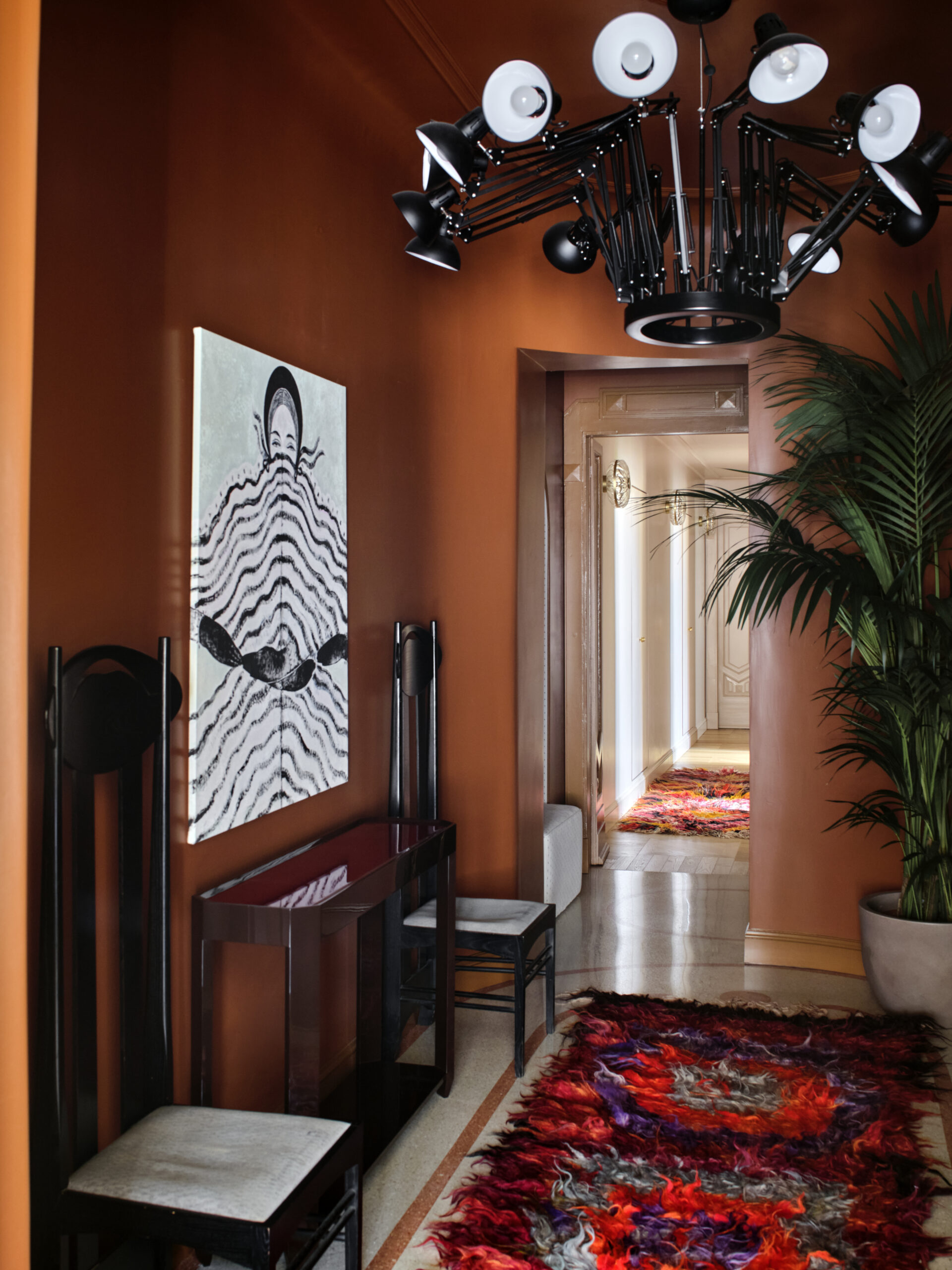 Disimpegno con pareti aranciate, console e sedie scure, lampadario a bracci e tappeto multicolore, aperto su un corridoio chiaro.