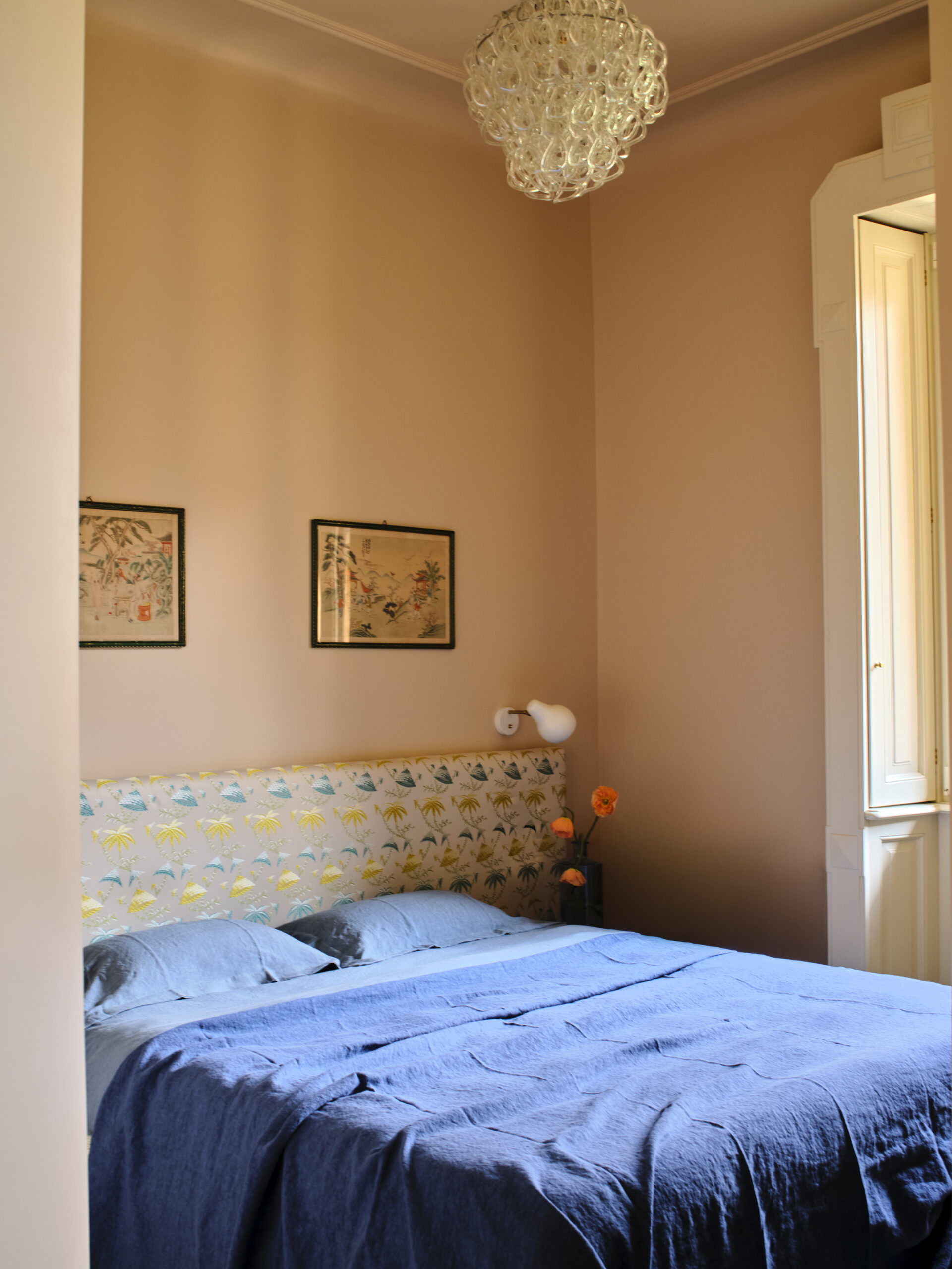 Camera con letto azzurro, testiera a motivi vegetali, pareti color pesca e lampadario trasparente vicino a una finestra.