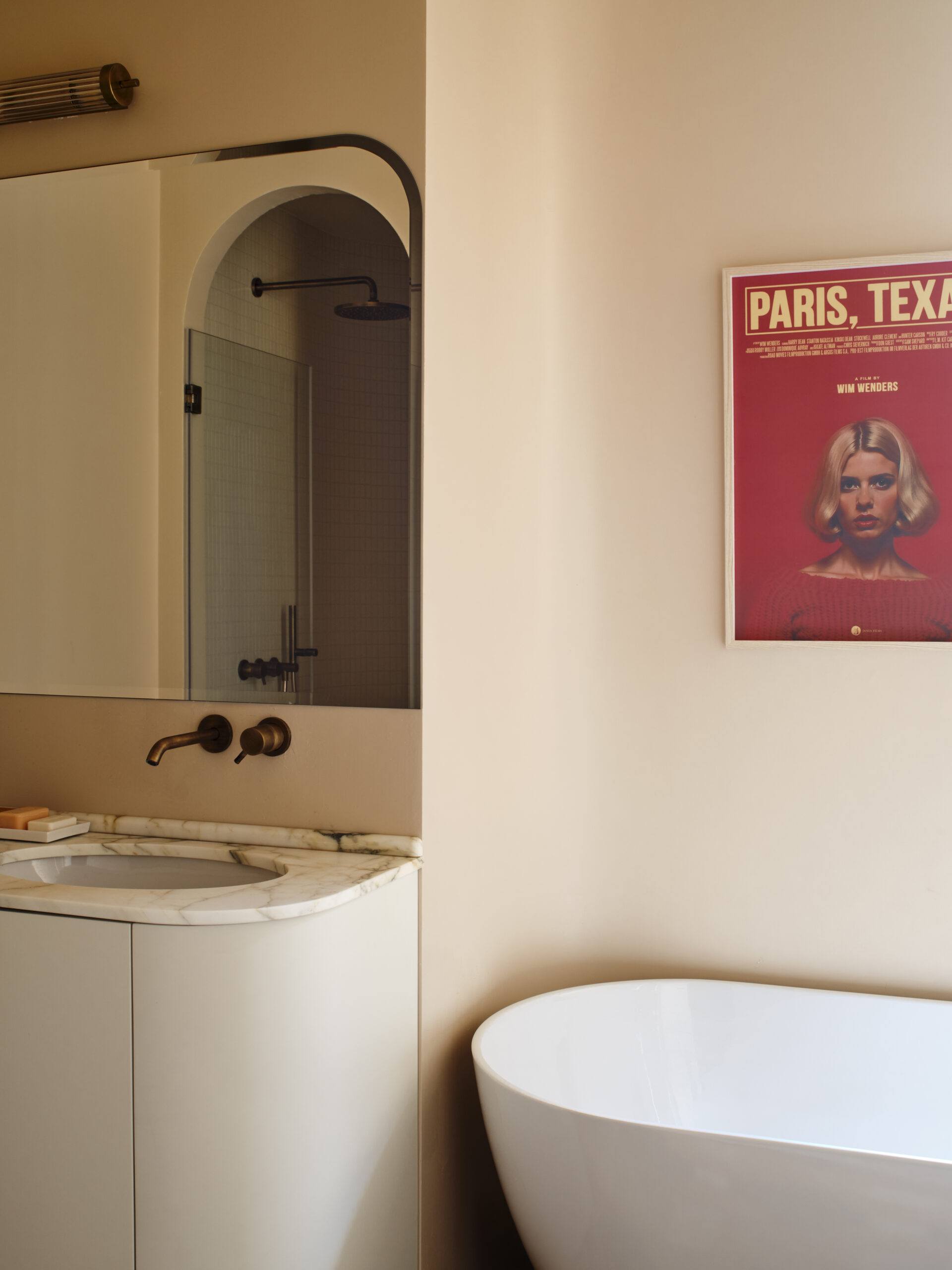 Bagno con lavabo su piano chiaro venato, specchio che riflette una doccia ad arco, vasca bianca e poster rosso sulla parete.