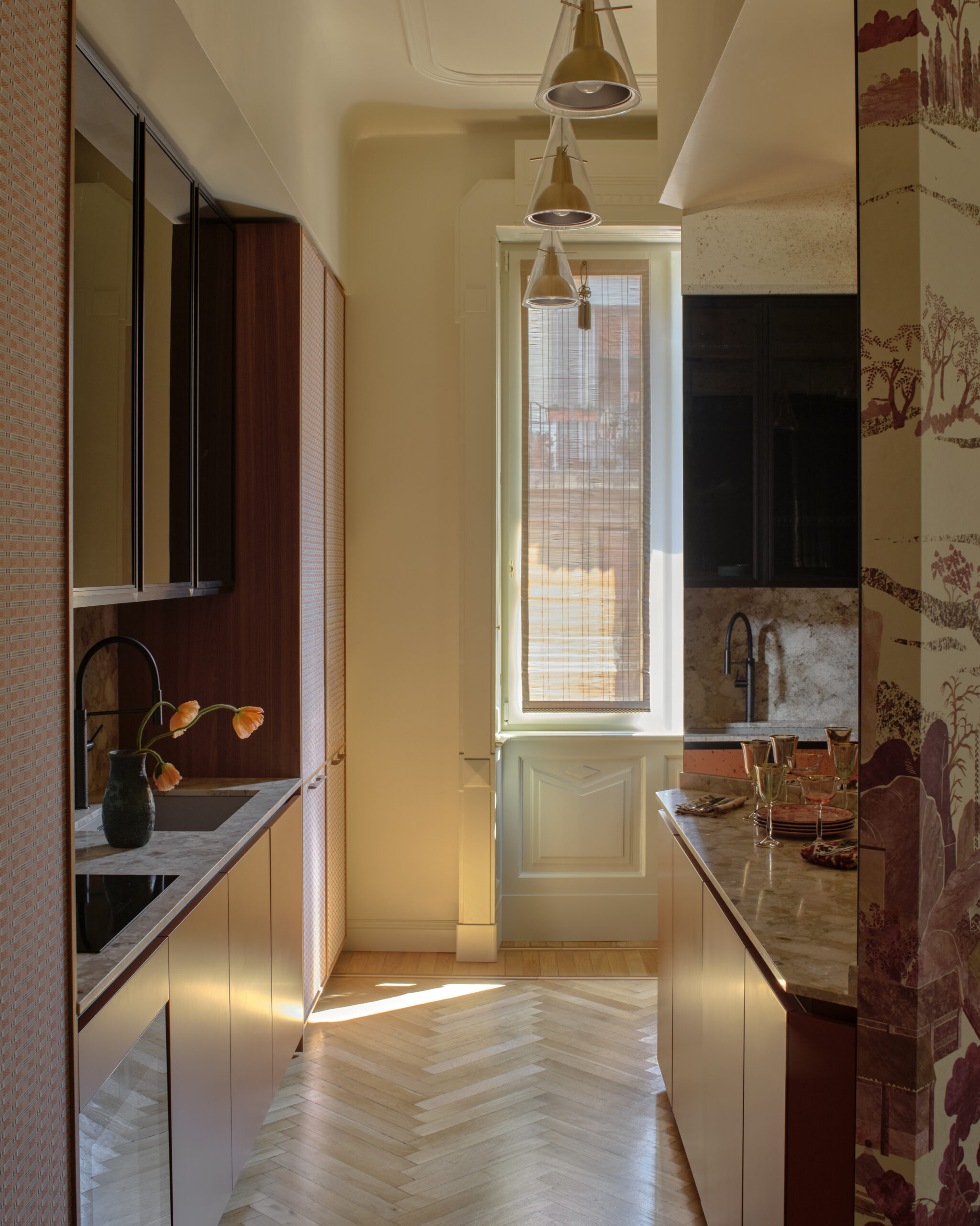 Cucina stretta illuminata da una finestra sul fondo, con ante color rame, piano chiaro, superfici riflettenti e tre lampade sospese.