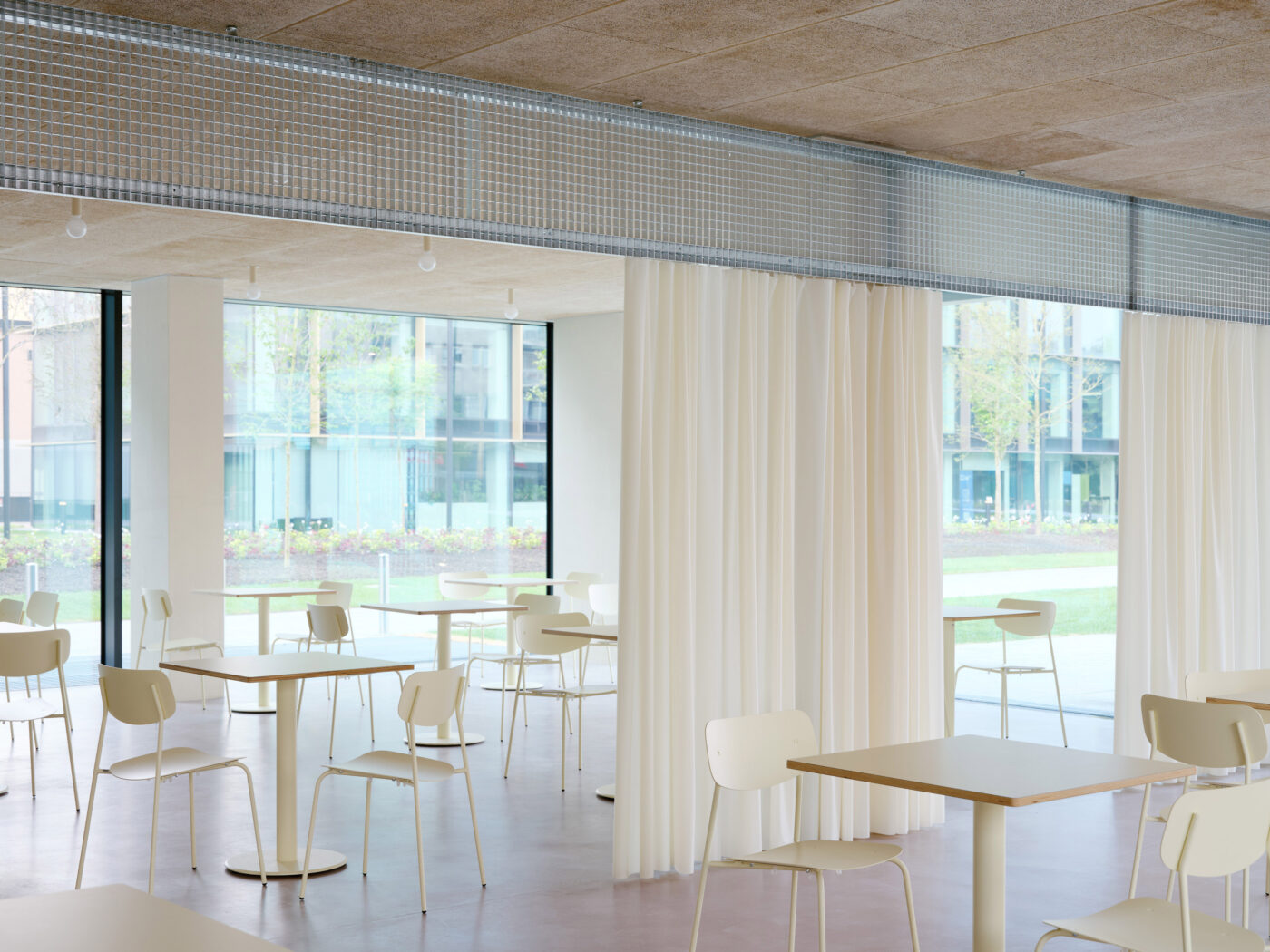 Sala luminosa con tavoli chiari, sedie sottili, tende color crema e fascia alta in policarbonato ondulato.