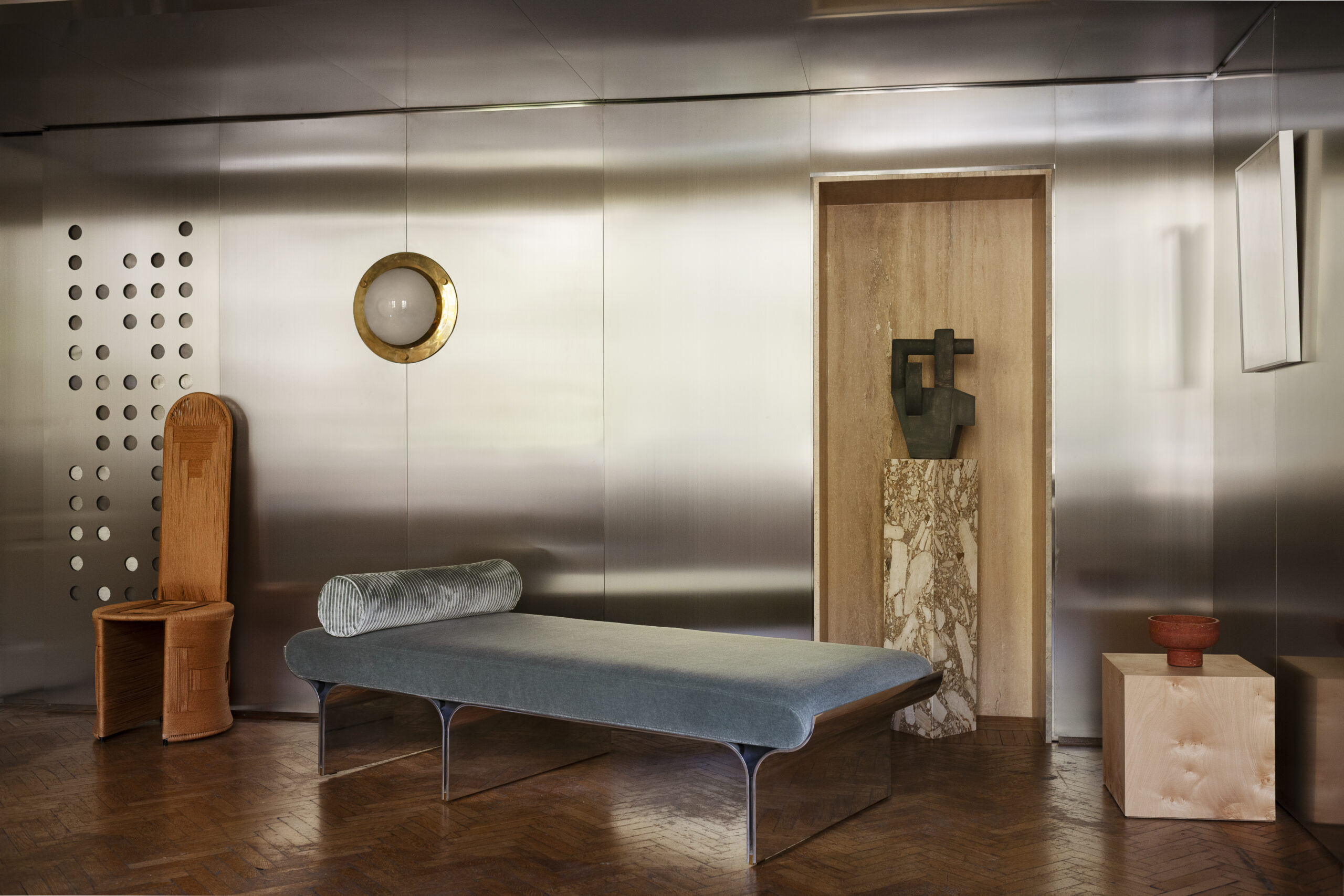 Daybed azzurro-grigio davanti a pareti metalliche riflettenti, con seduta alta a sinistra e scultura scura su piedistallo a destra.
