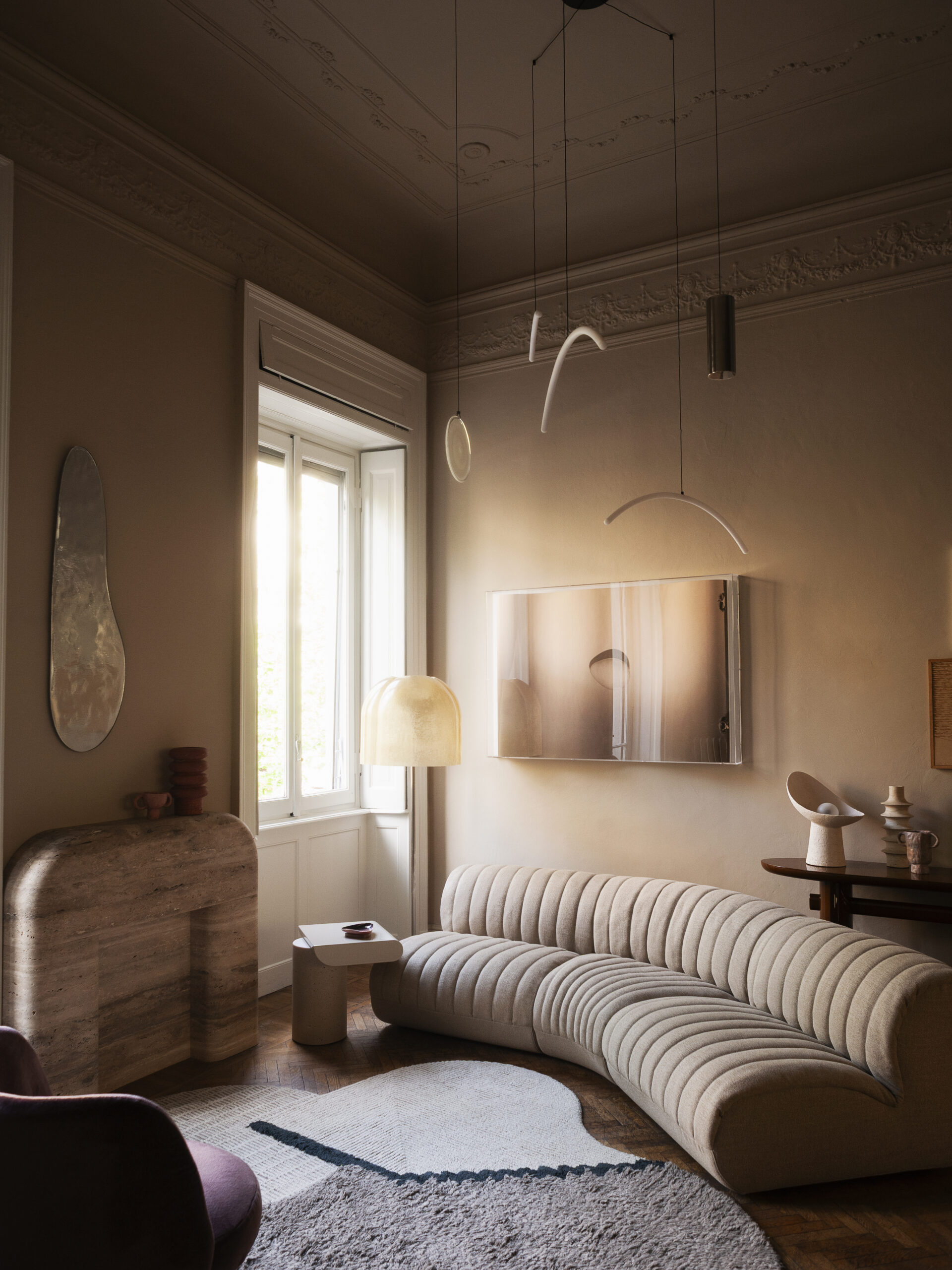 Divano chiaro curvo e scanalato sotto una composizione sospesa, accanto a un elemento in pietra arrotondato; soffitto decorato e tappeto sagomato.