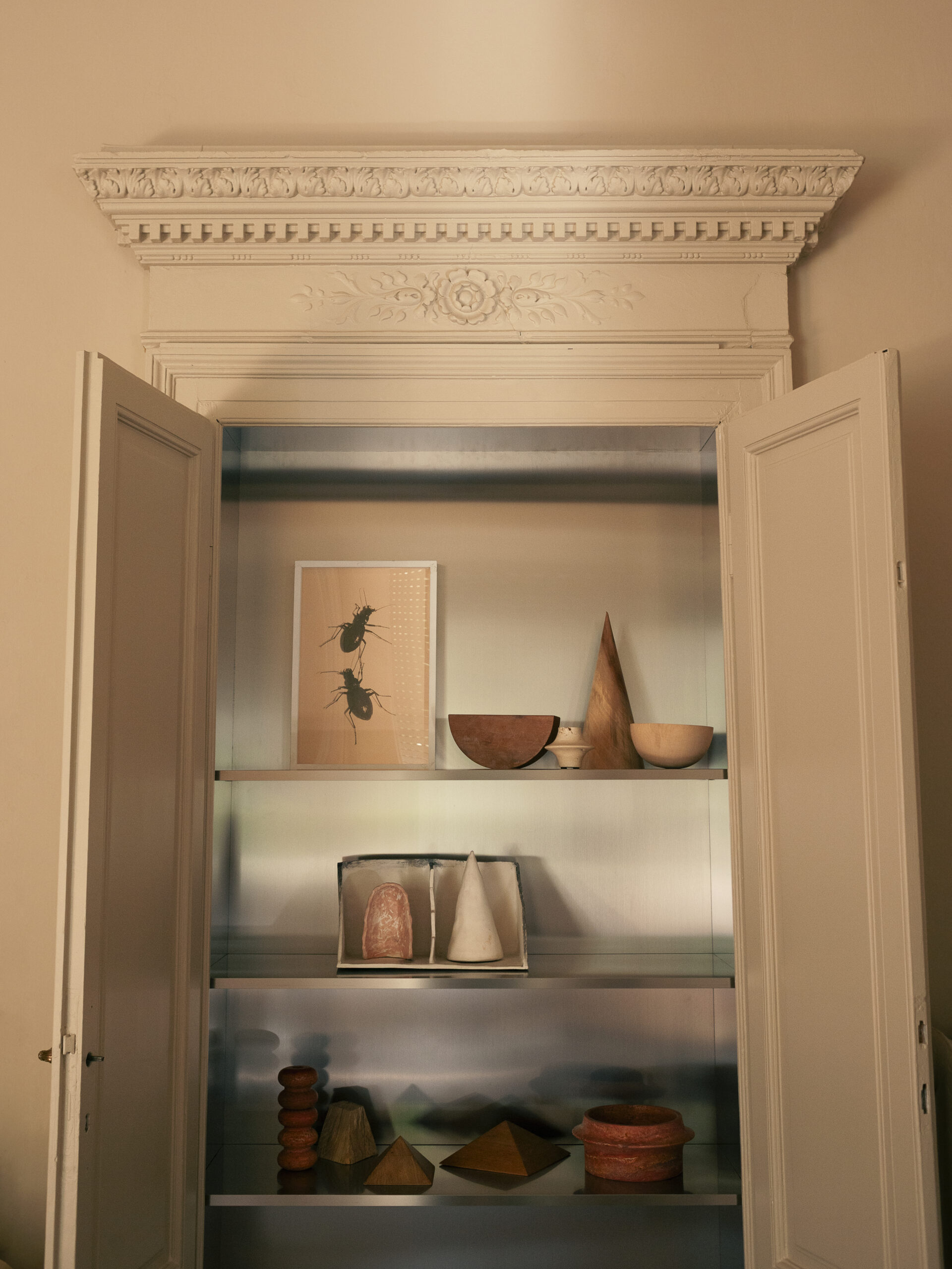 Nicchia con ripiani metallici, ciotole, forme geometriche e un quadro con insetti, fra due ante chiare sotto una cornice decorata.