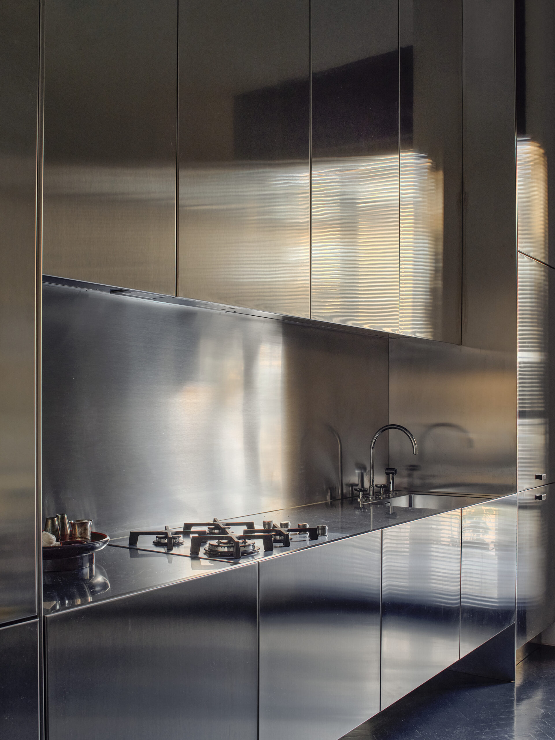 Cucina lineare con ante, piano e alzata metallici, fornelli a gas e lavello attraversati da riflessi di luce.