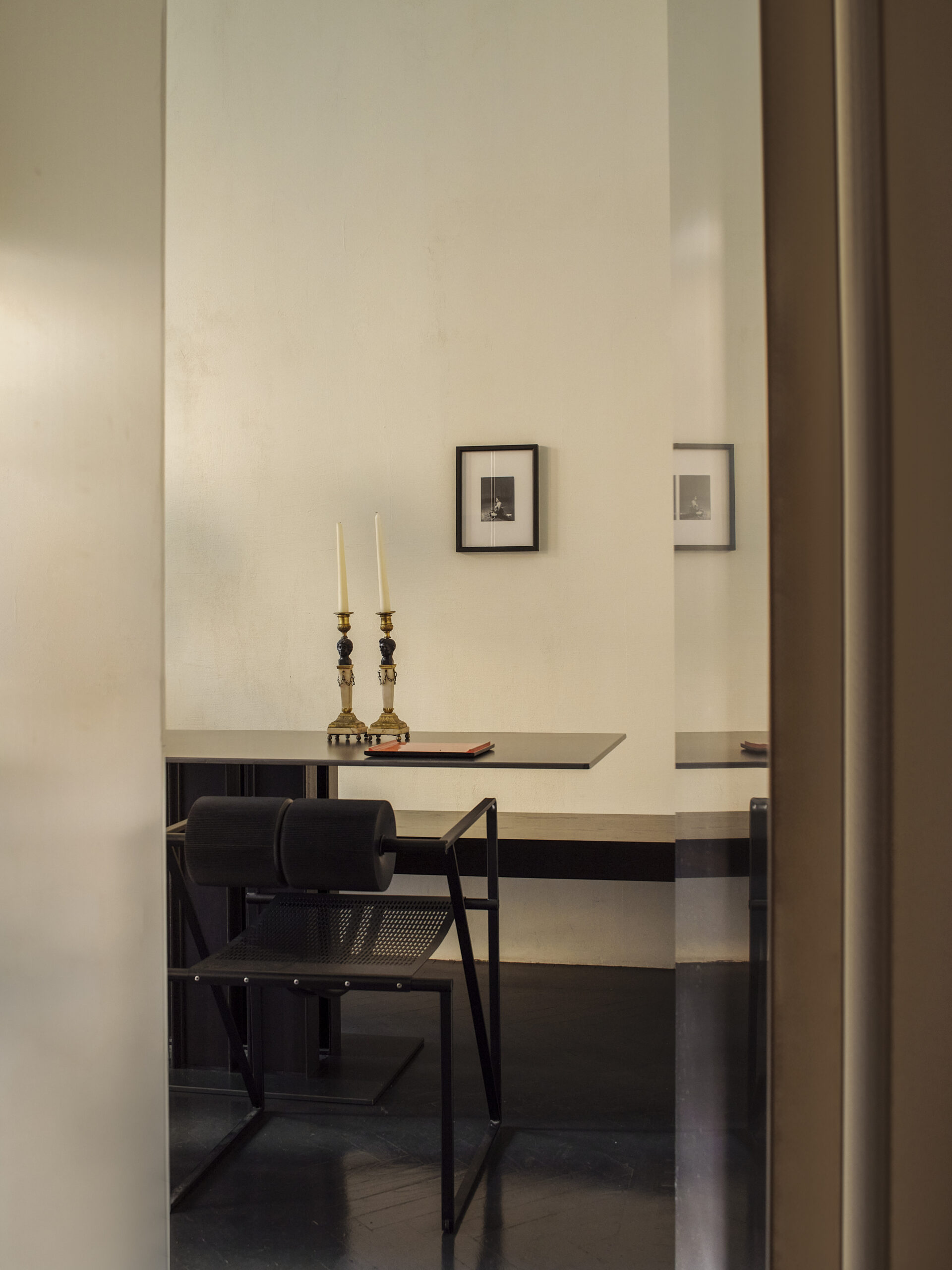 Tavolo sottile con due candelabri, una sedia nera e una fotografia incorniciata, visto attraverso un'apertura con un pannello riflettente.