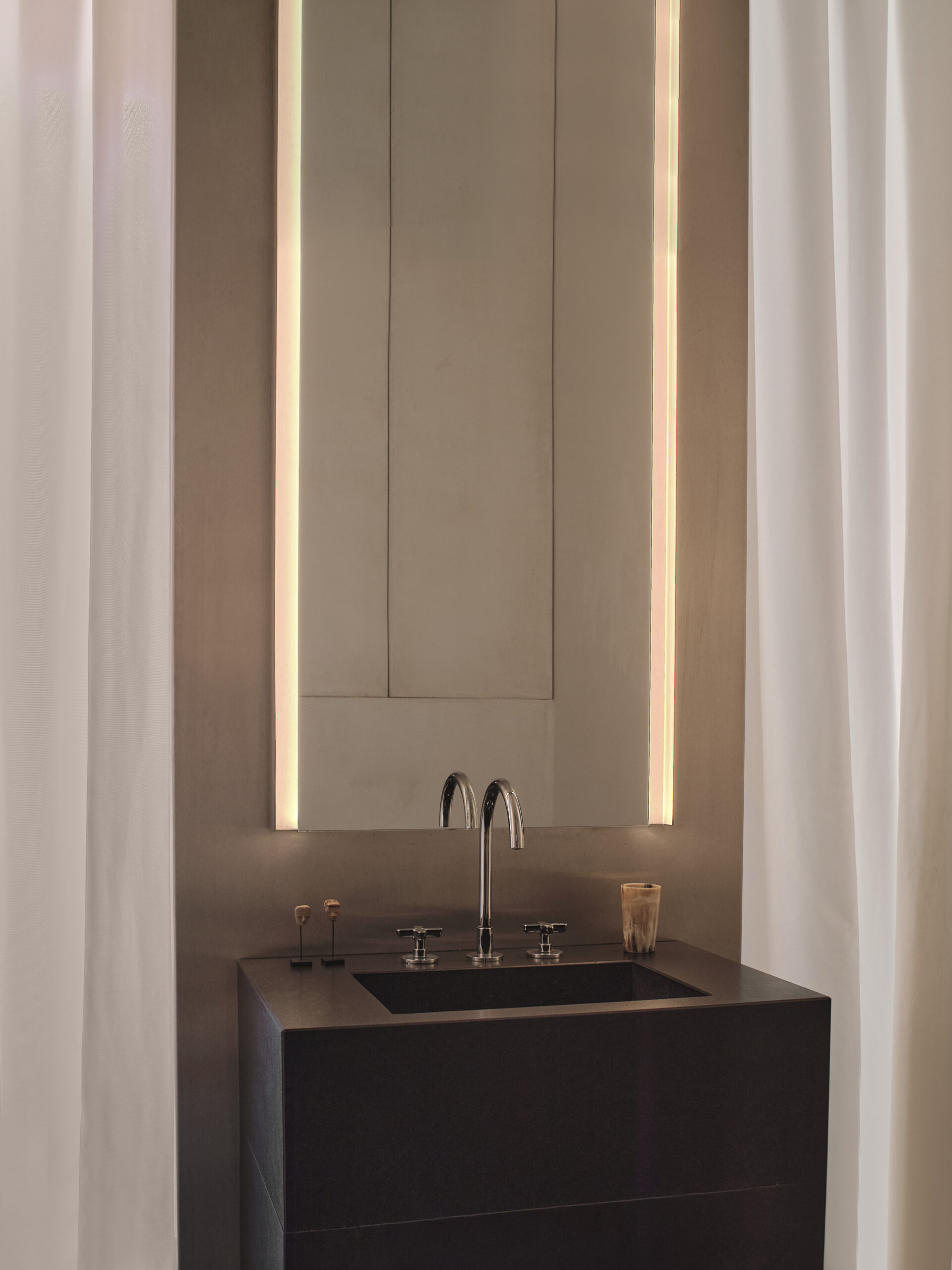 Lavabo scuro con rubinetteria cromata sotto uno specchio alto illuminato ai lati, tra tende chiare.
