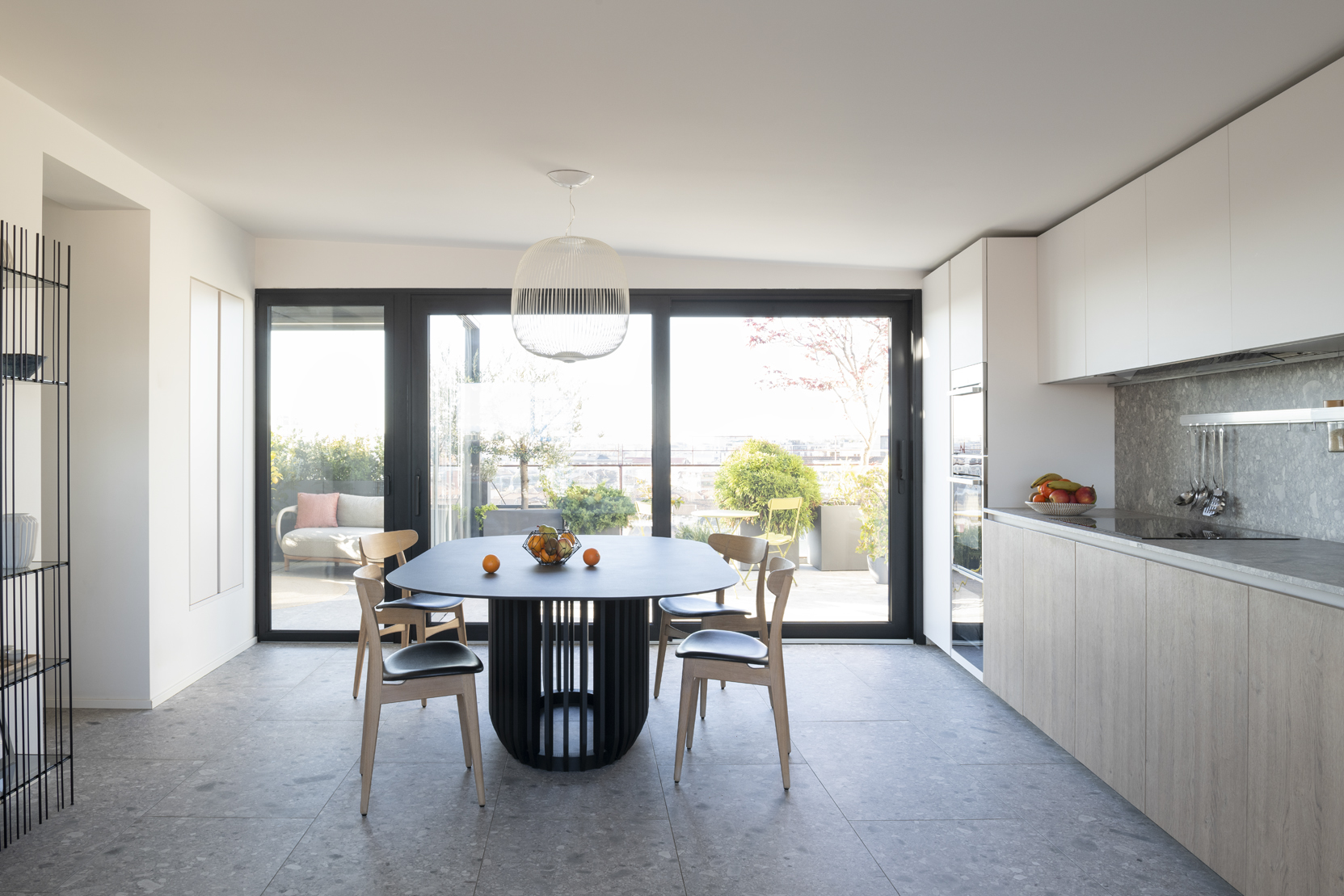 Cucina e zona pranzo con tavolo scuro, sedie in legno e ampie vetrate aperte visivamente sul terrazzo.