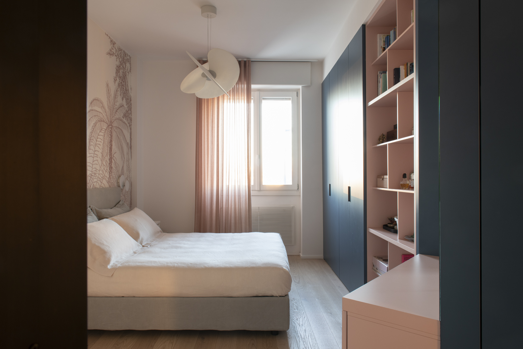 Camera con letto chiaro, armadio blu, libreria cipria, tende rosa e carta da parati botanica.