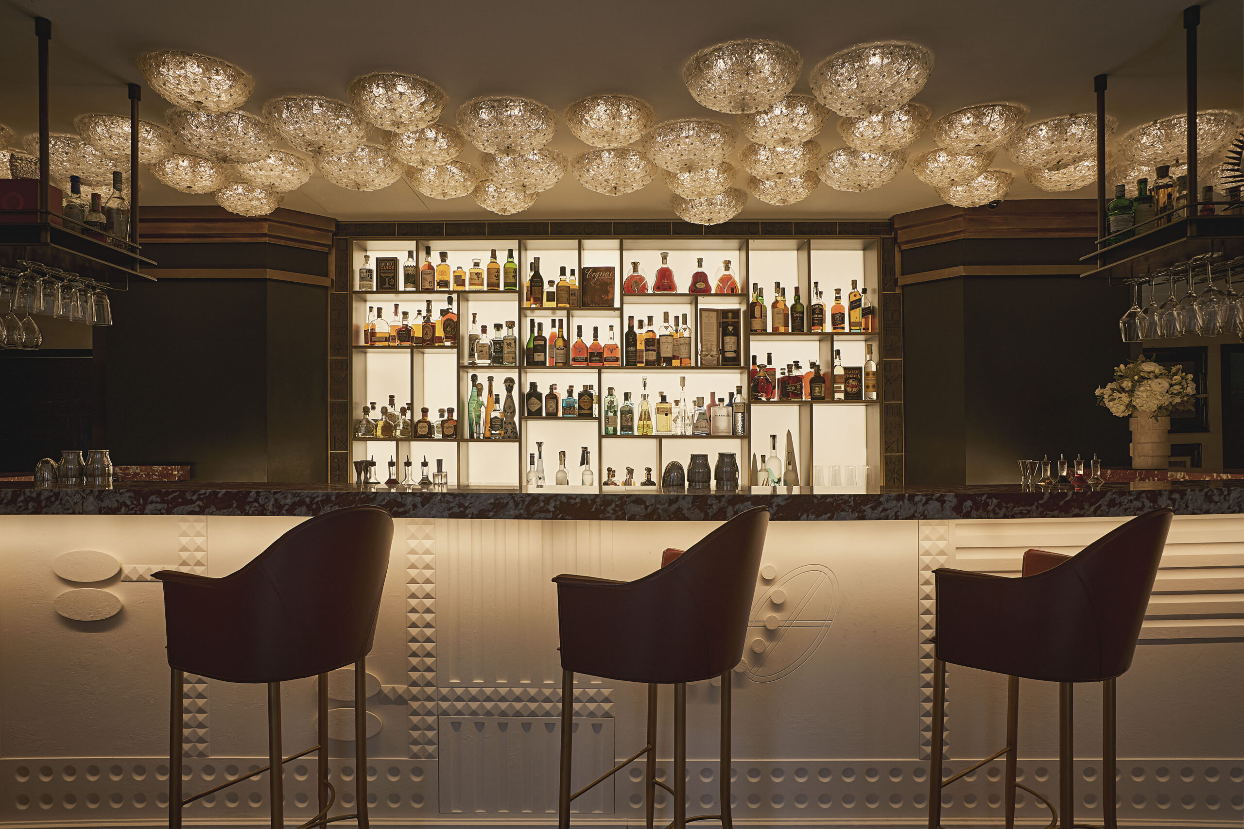Carlton Bar con plafoniere in vetro, bottigliera illuminata, bancone a rilievi geometrici e sgabelli scuri.