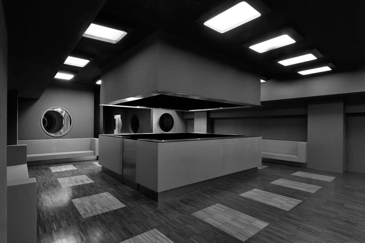 Fotografia in bianco e nero del bar, con bancone geometrico, pavimento a listelli, aperture circolari e plafoniere quadrate.