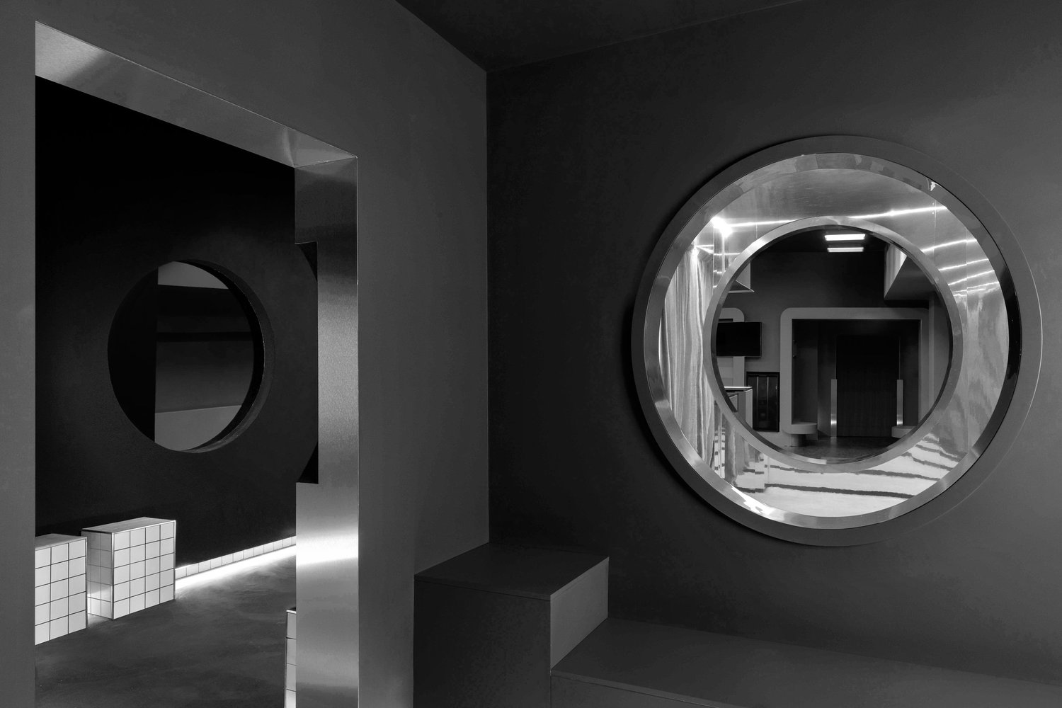 Interno in bianco e nero con aperture circolari, pareti scure, un oblò metallico e un volume basso rivestito a quadretti.