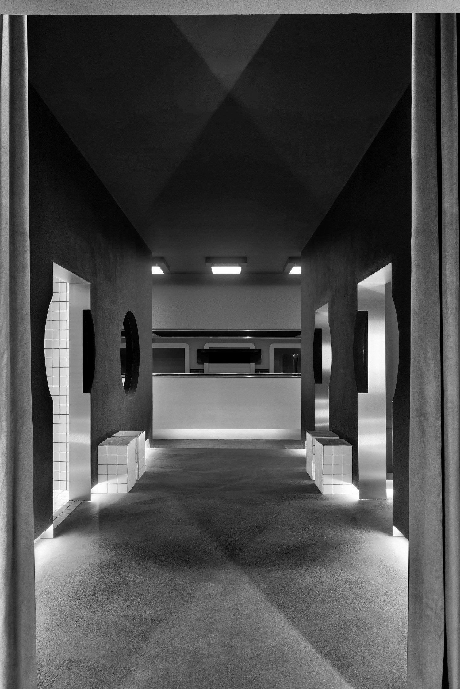 Corridoio in bianco e nero con pareti scure, aperture laterali e bancone sul fondo sotto plafoniere quadrate.