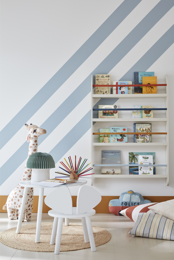 Children's reading corner with muted blue and white wall stripes, bookshelves, pale furniture and a woven rug