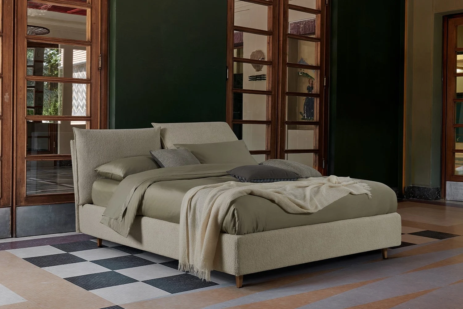 Beige upholstered bed with a soft headboard in a bedroom with dark green walls and a chequered floor