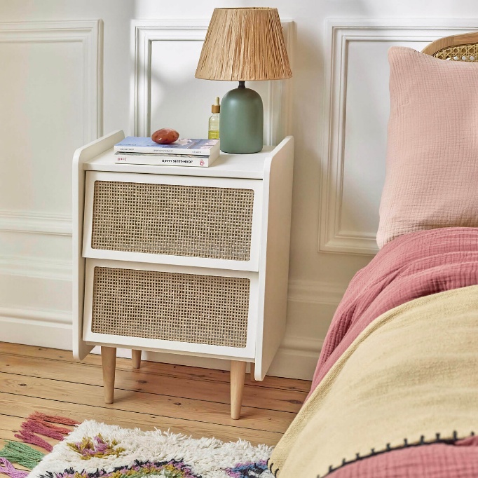Comodino bianco con due cassetti in rattan, lampada da tavolo e letto con tessili rosa e beige.