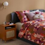 Comodino color terracotta con anta in rattan accanto a un letto imbottito con biancheria floreale nei toni del rosa e del bordeaux.