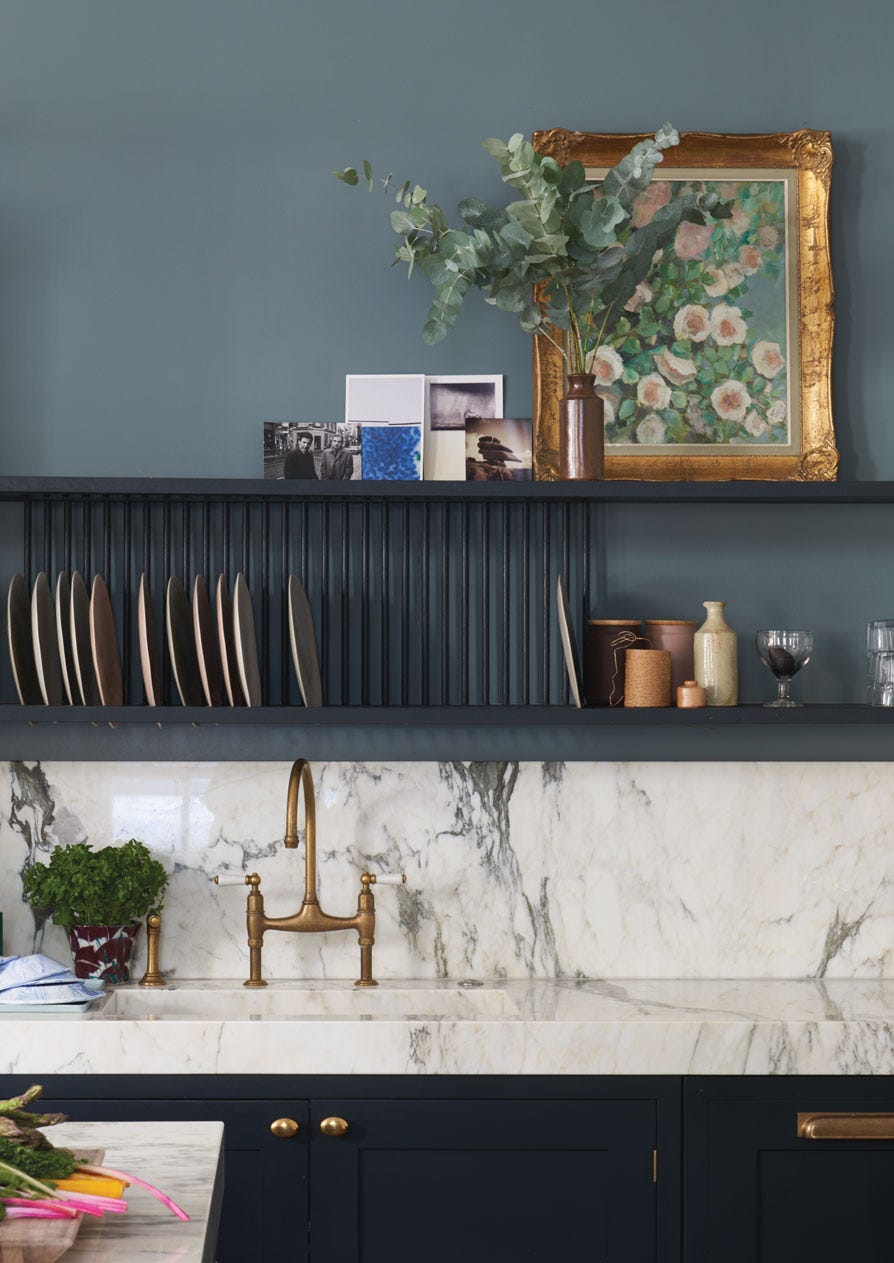 Kitchen with a grey-blue wall, darker cabinetry, white veined surfaces and a brass tap