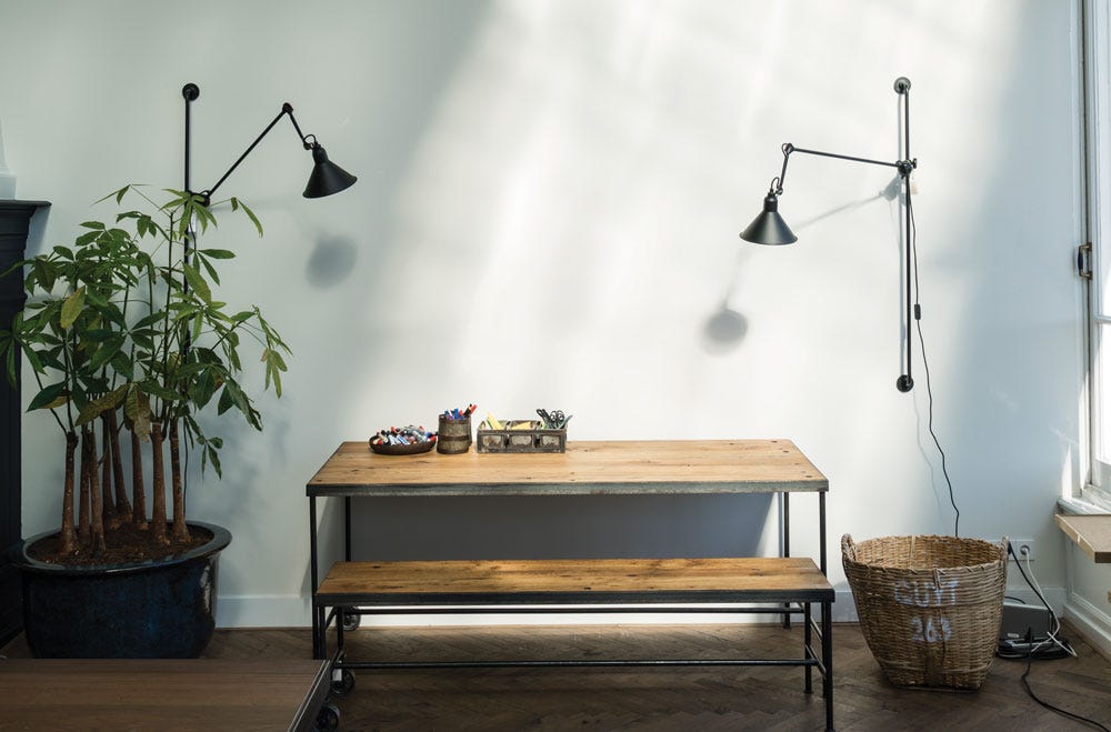 Daylight and shadows on a pale cool wall, with a timber table and bench, black lamps and a plant beside the window