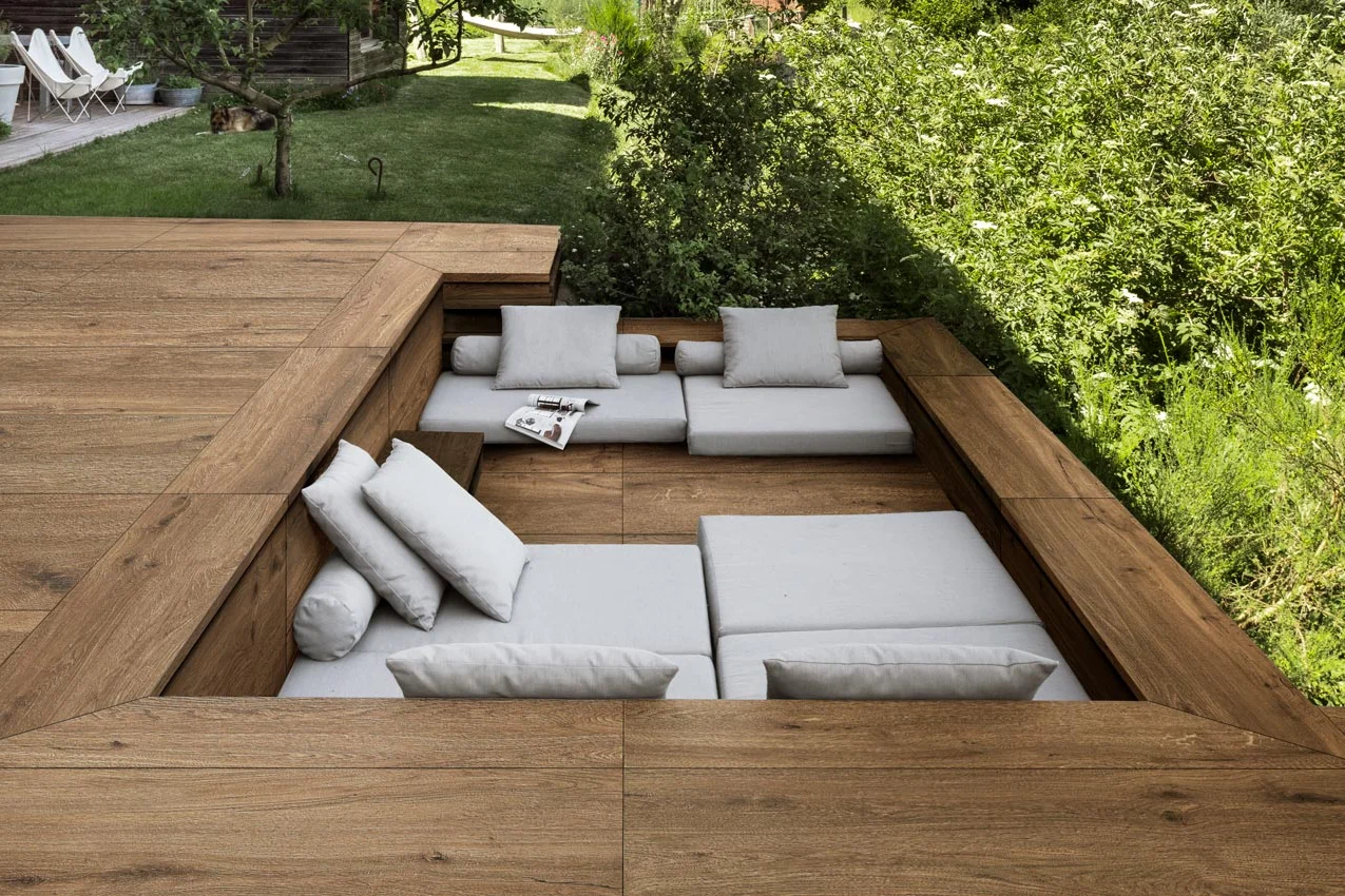 Salotto da esterno ribassato nel giardino con pavimento e sedute rivestite in gres effetto legno spessorato